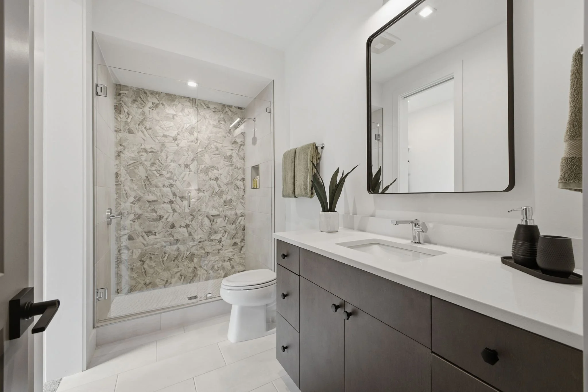 Modern bathroom with a walk-in shower featuring marble tiles, a white vanity with a rectangular black-framed mirror, and a potted plant on the counter.