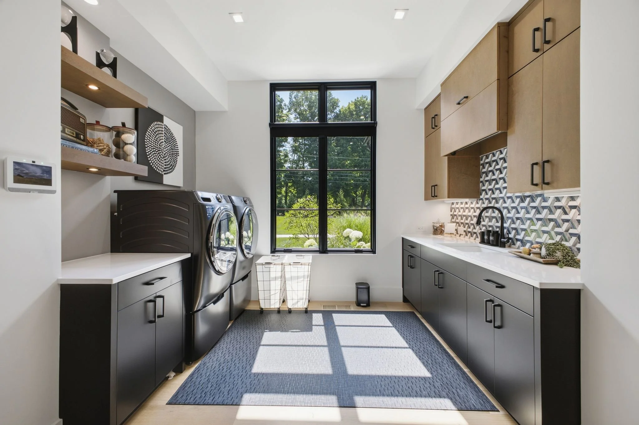 Modern laundry room with black washer and dryer, white countertops, wooden upper cabinets, geometric backsplash, large window, and decorative items.