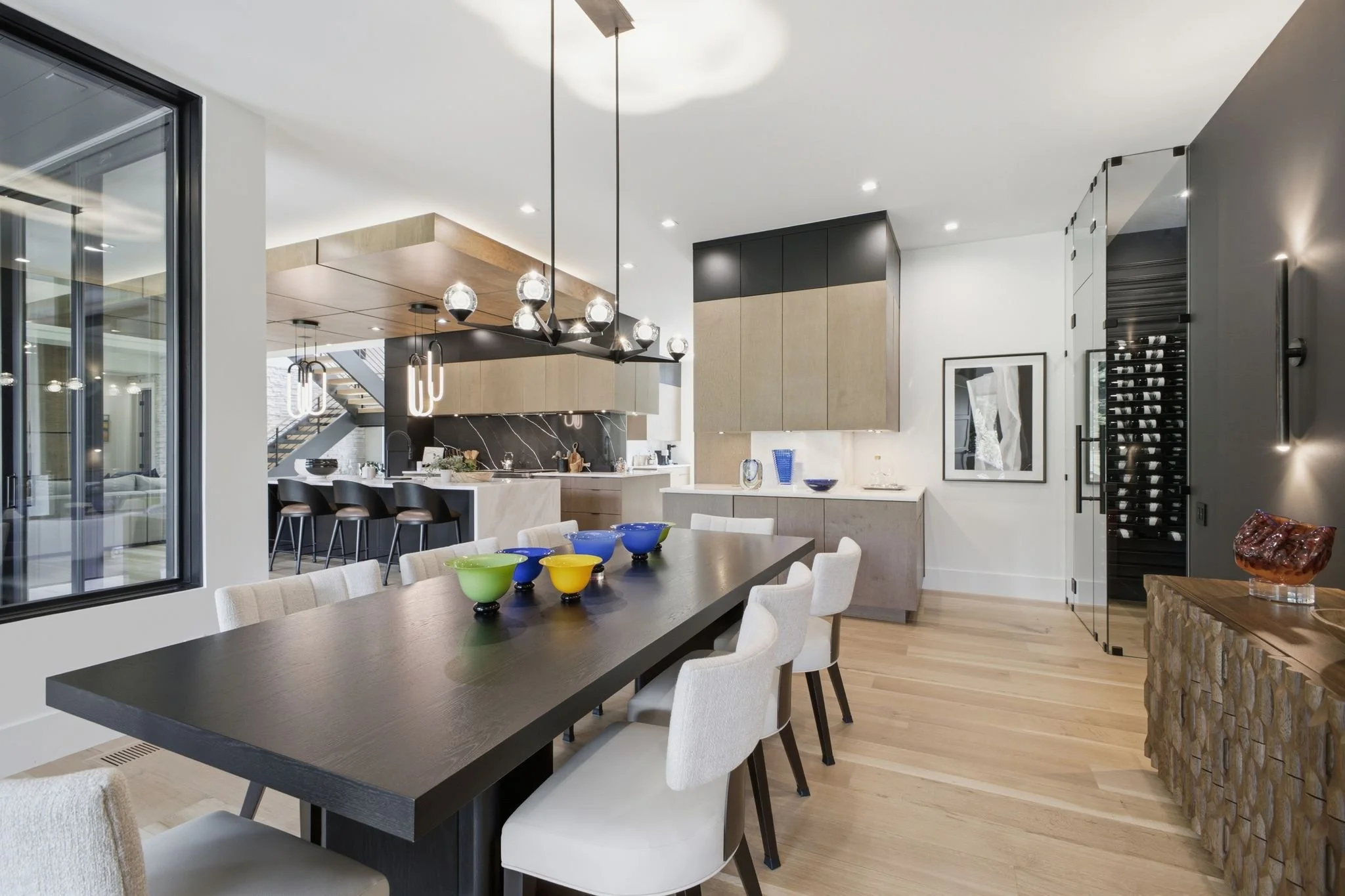 Modern open-concept kitchen and dining area with black dining table, white upholstered chairs, colorful bowls, pendant and chandelier lighting, wooden and black cabinetry, wine storage, and decorative art.