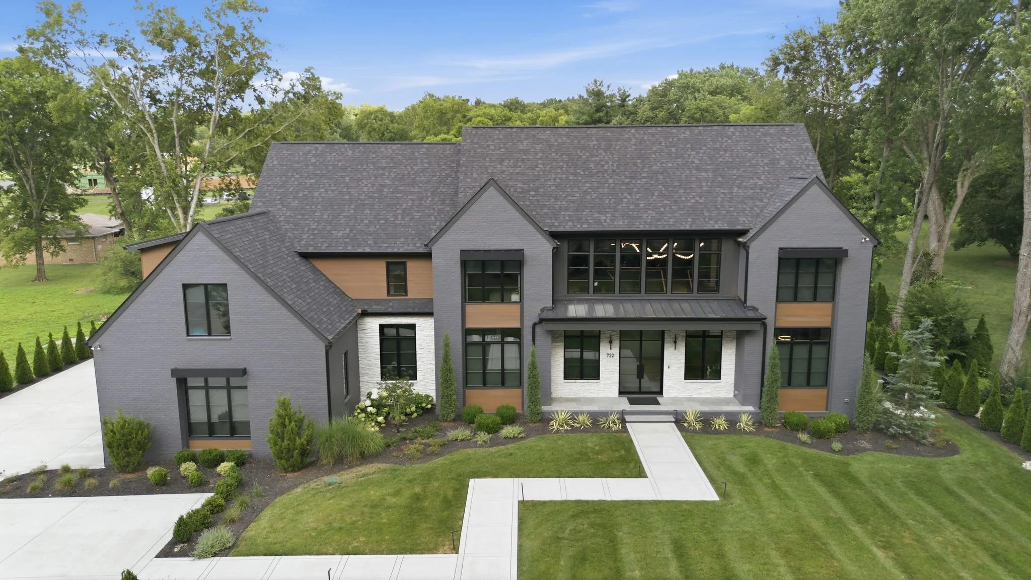 A modern two-story house painted in gray with black window frames and accents, a white brick front entrance, and a landscaped front yard with walkway and greenery, surrounded by tall trees under a blue sky.