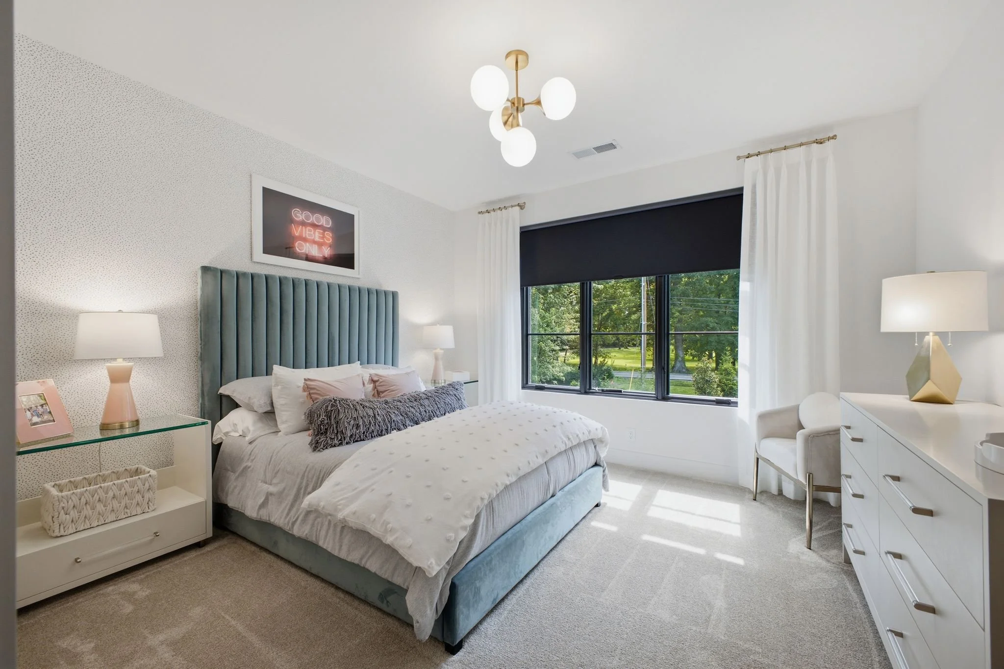 A modern bedroom with a large bed featuring a tall, teal, upholstered headboard, white bedding, and decorative pillows. There are twin white nightstands with lamps on each side of the bed, a framed neon sign that reads 'Good Vibes Only' above the hea