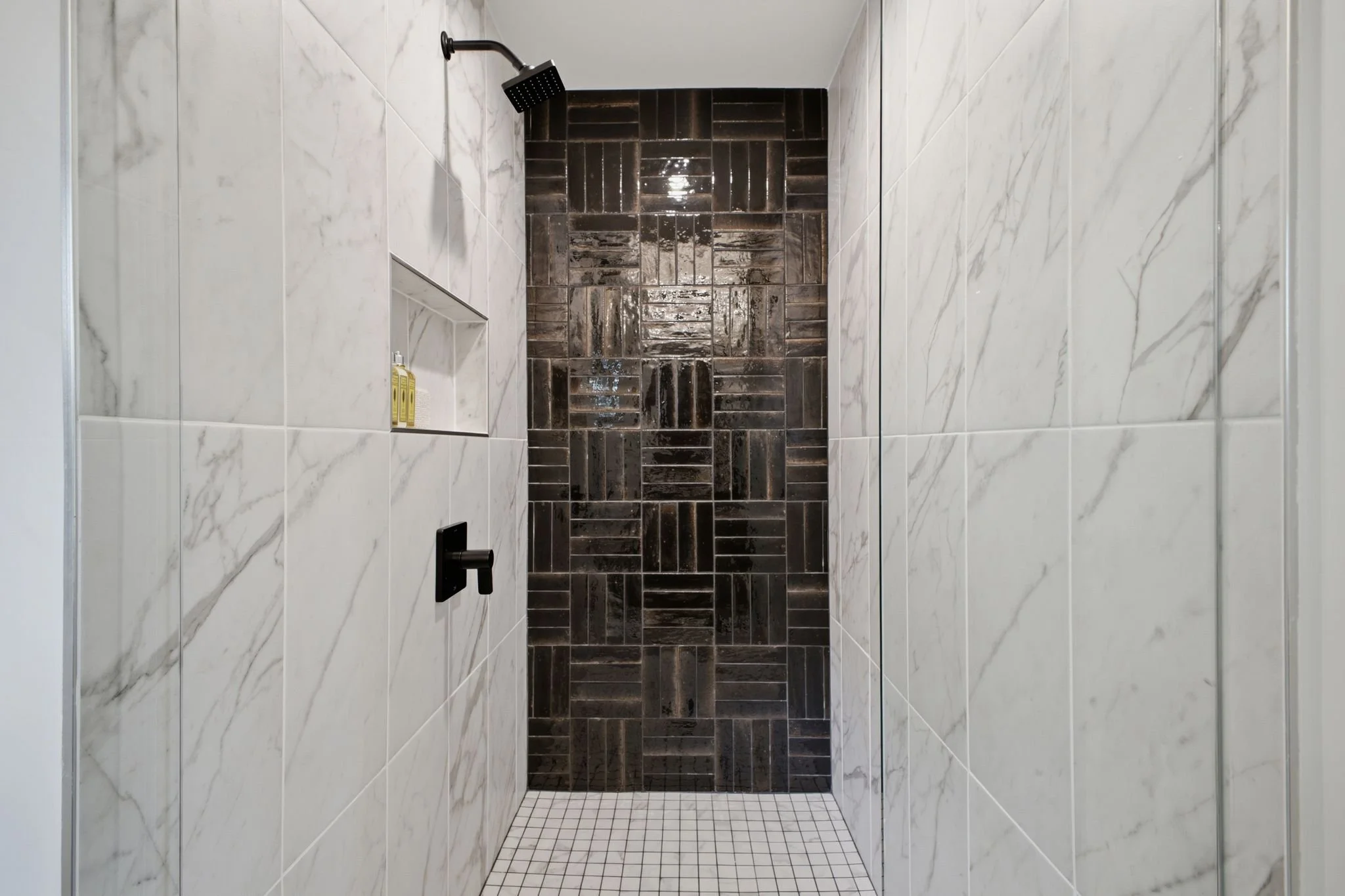 A shower with white marble tiles on the walls, a dark tiled accent wall, a black showerhead, and a built-in shelf with a yellow soap bottle.