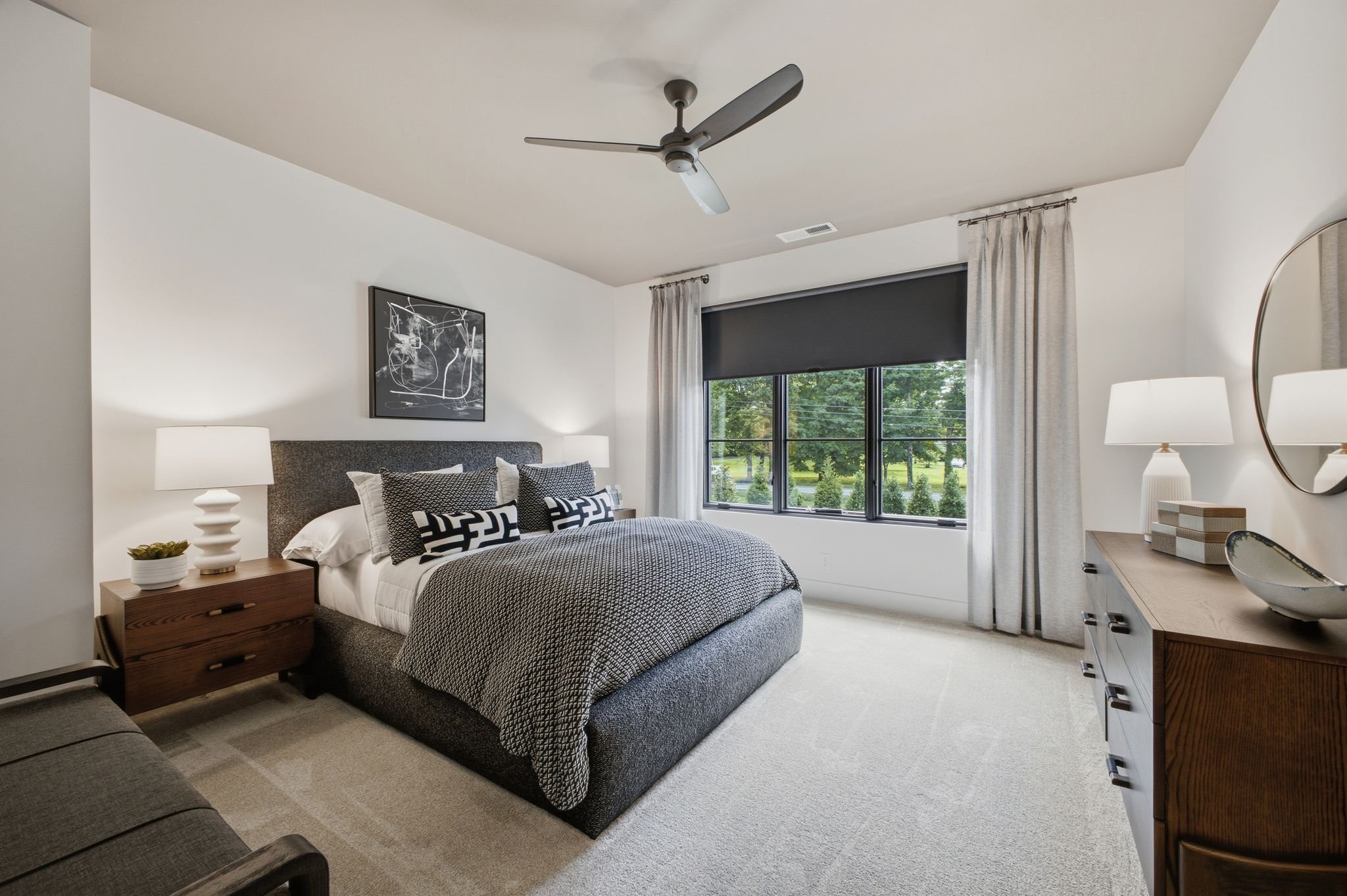 A modern bedroom with a bed, nightstand, lamps, window with curtains, and wall art.