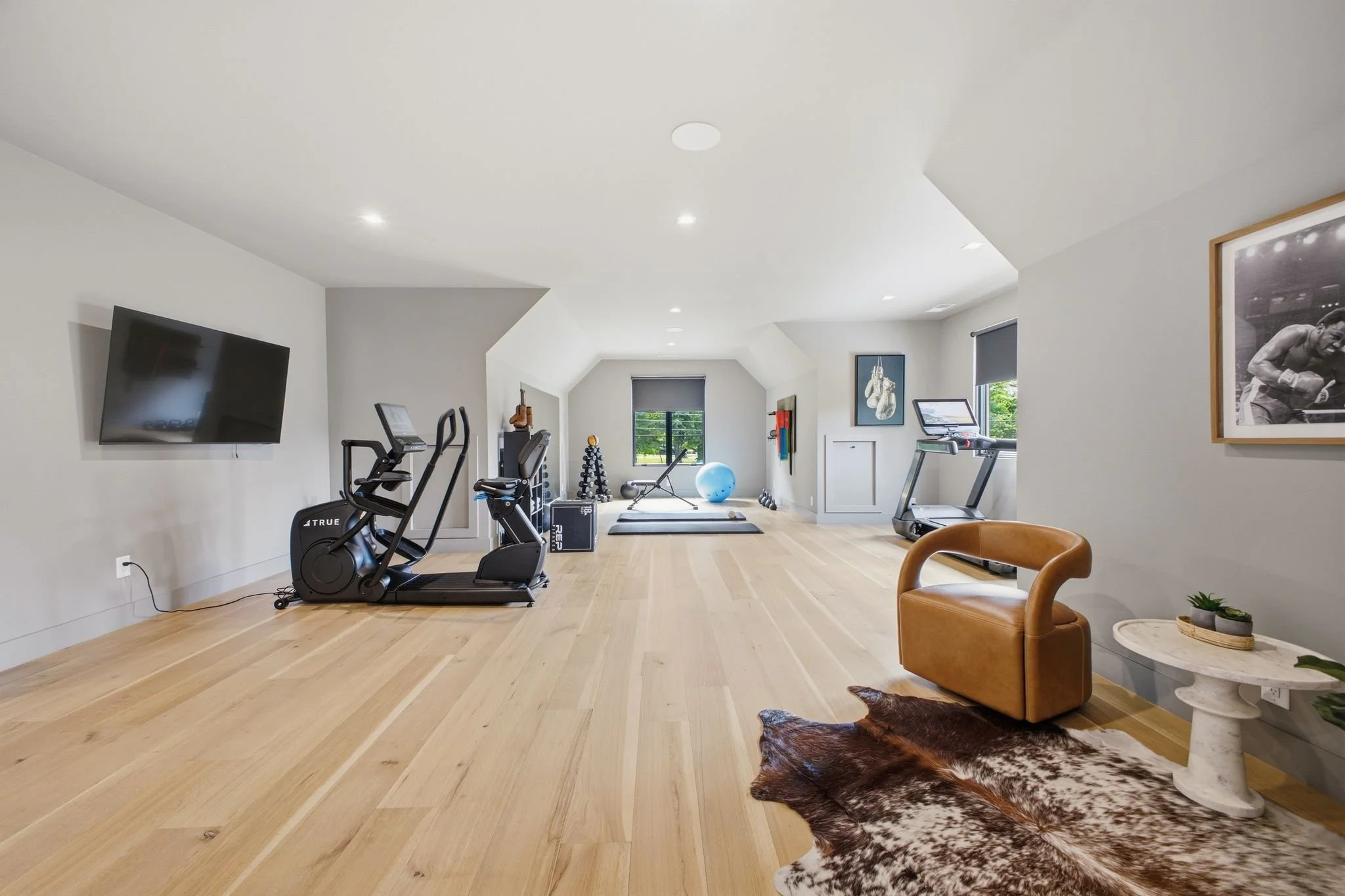 Home gym with a wall-mounted TV, exercise bike, elliptical machine, treadmill, and free weights, all in a bright room with hardwood floors and a large window.
