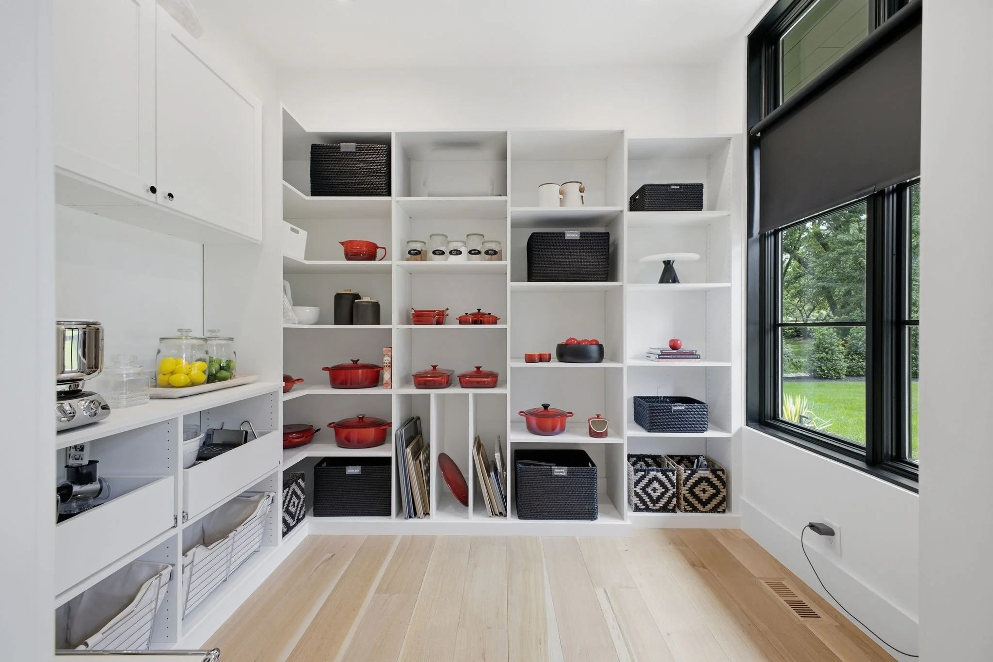 A bright kitchen with white shelving filled with black and red cookware, glass jars, baskets, and framed artwork, near large windows with black blinds overlooking a green yard.