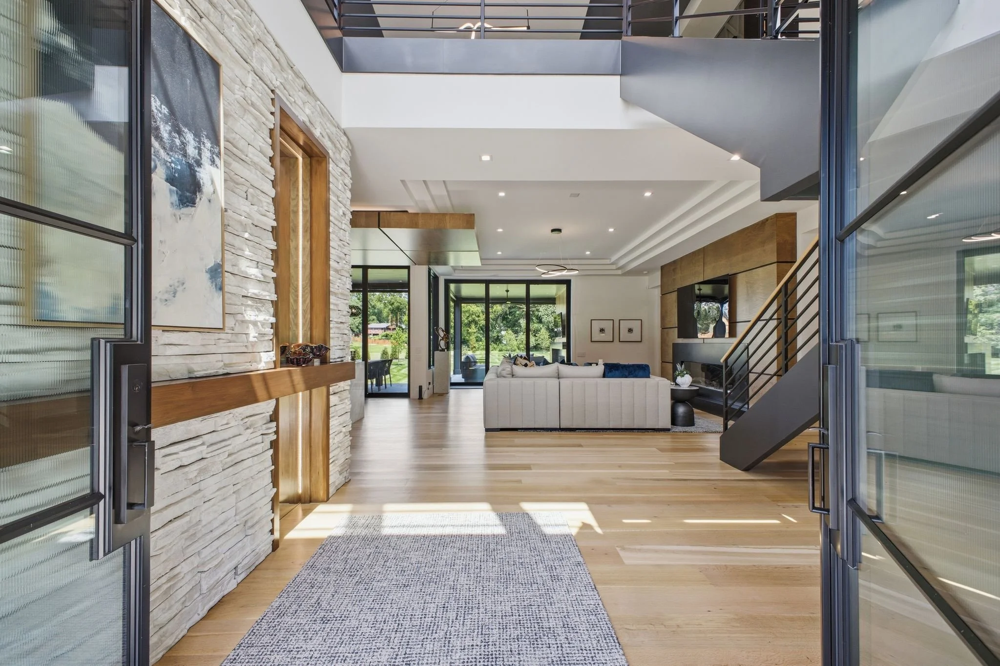 View of a spacious, modern living room inside a house, seen through open glass doors. The room has a stone accent wall, wooden accents, recessed ceiling lights, and large windows showcasing greenery outside. Furniture includes a cream-colored section