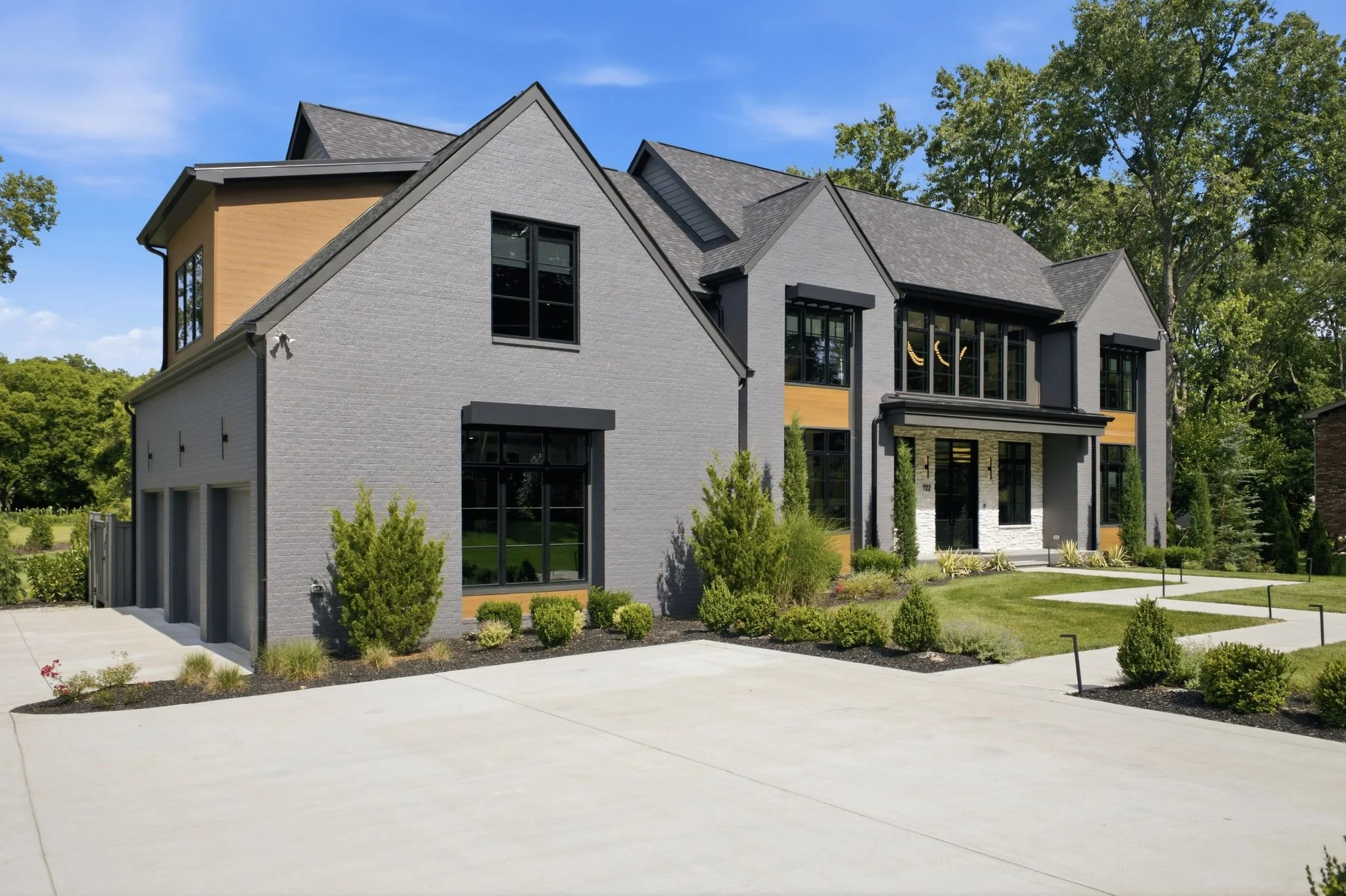 Modern two-story house with gray brick exterior, large windows, and landscaped front yard with small bushes and grass, under a blue sky with some clouds.