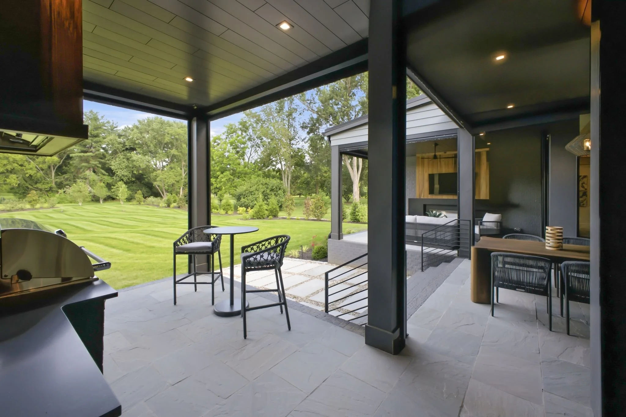 Modern patio with a black metal chair and table, outdoor grill, and seating area with a view of a lush green yard with trees and grass.