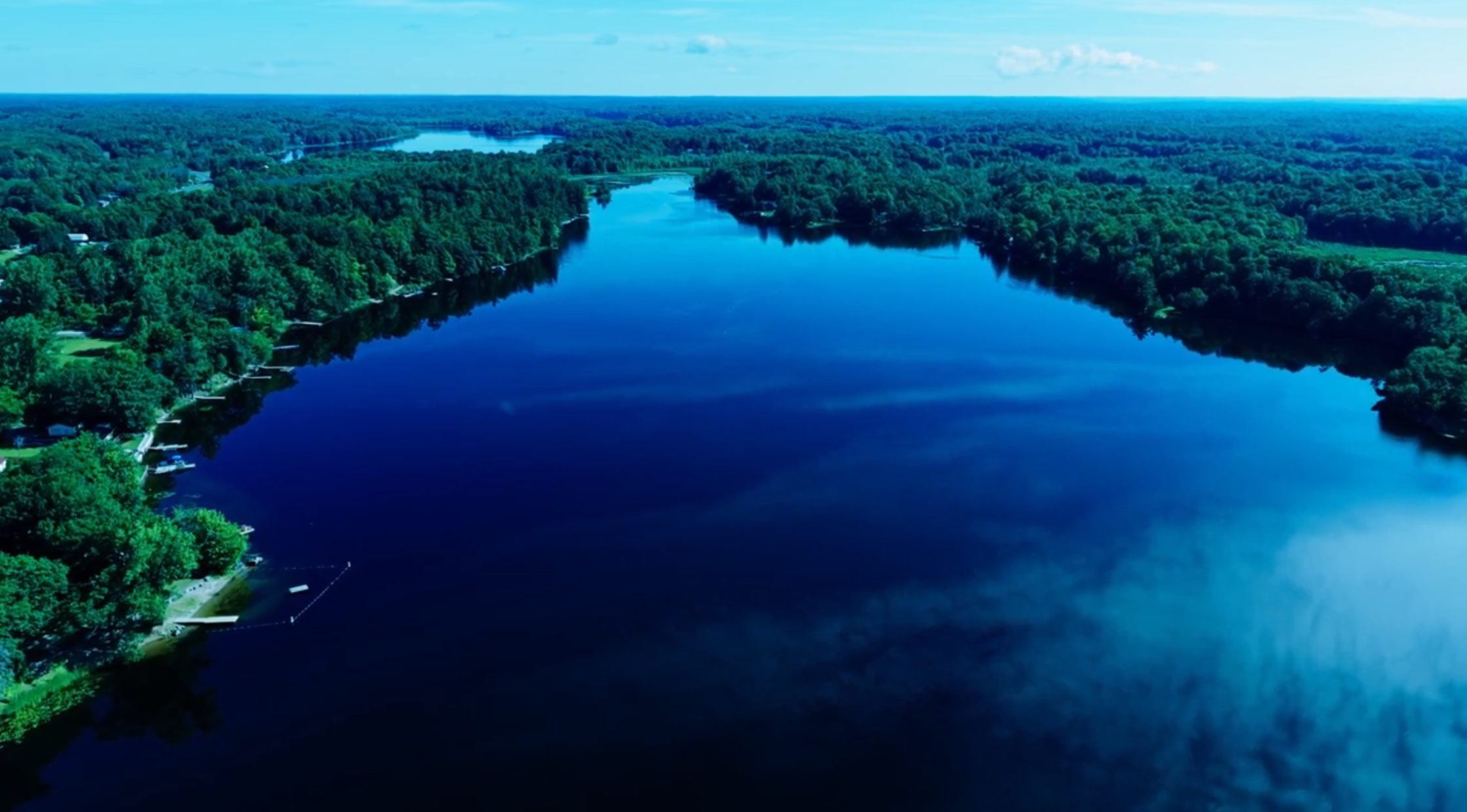 Aerial view of a river winding through a lush green forest with a few houses along the shoreline under a partly cloudy sky.