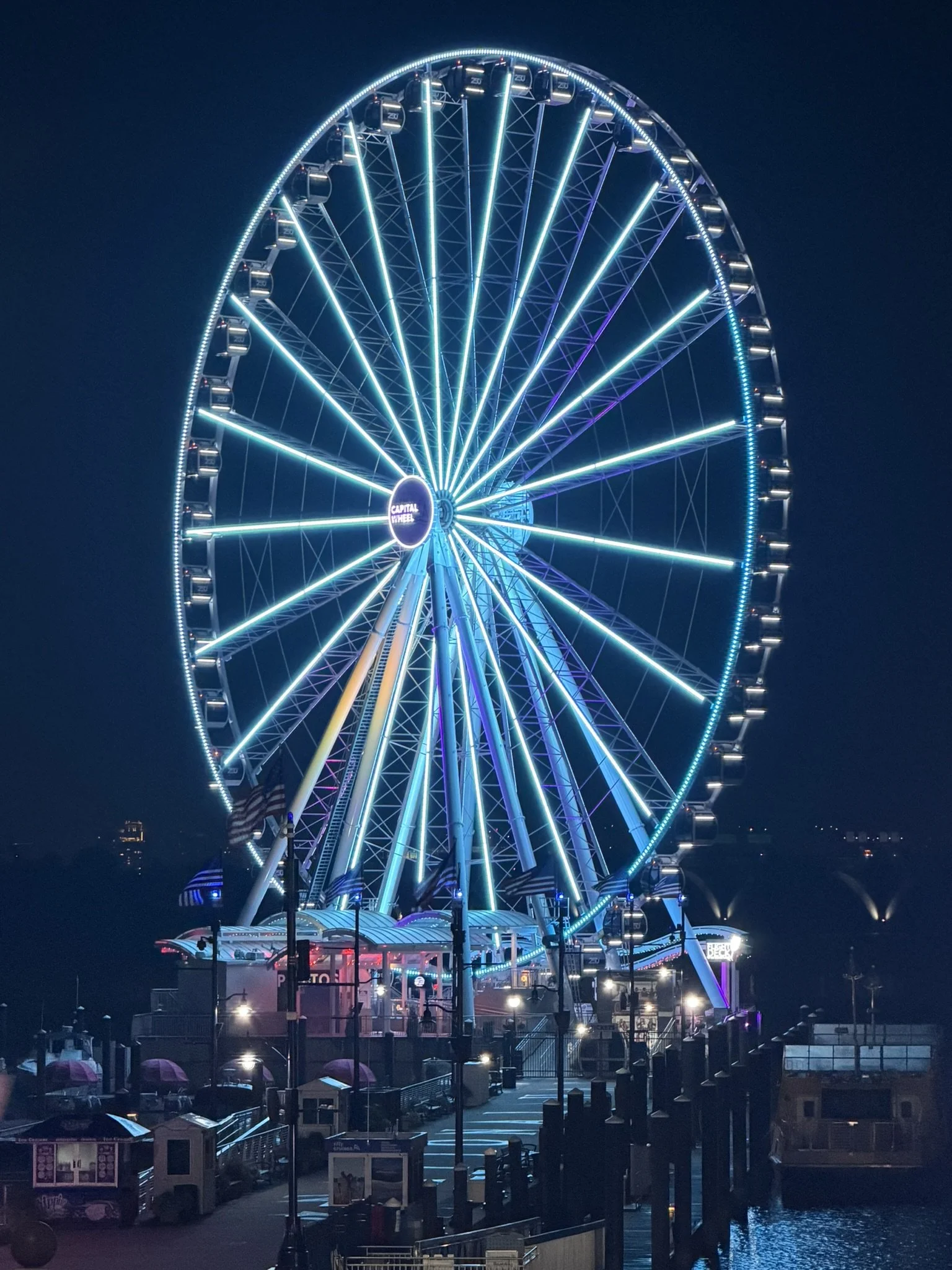A large illuminated Ferris wheel at night, featuring bright blue and green lights, with a small arcade and ticket booth area in the foreground by the water.