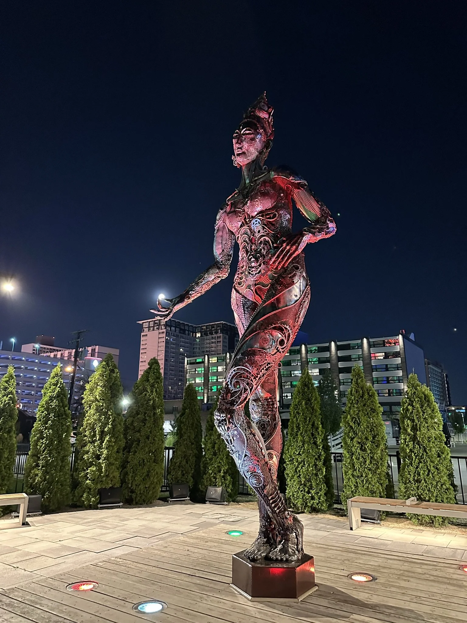 Nighttime cityscape featuring a tall, modern sculpture of a human figure with intricate, metallic, and illuminated surfaces, surrounded by a row of tall green pine trees, with multi-story buildings in the background and colorful lights on the ground.