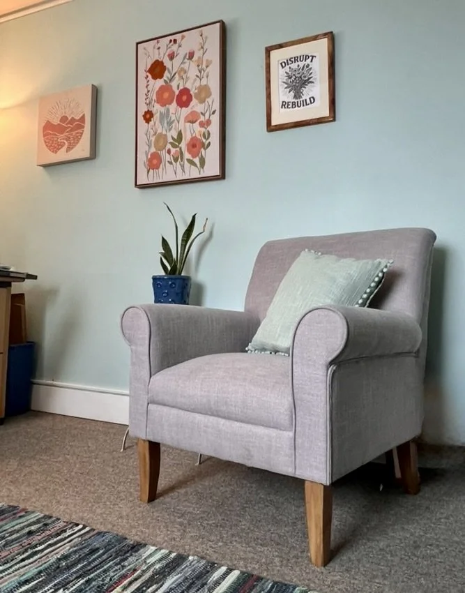 A cozy living room corner with a light gray armchair with wooden legs, a light green cushion on it, and a potted plant in a blue ceramic pot on a small table next to it. The wall behind has three framed artworks: a heart-shaped landscape, a colorful floral print, and a black and white print with the words 'DISRUPT REBUILD' and a leaf illustration. A textured rug with multicolored stripes is on the floor.