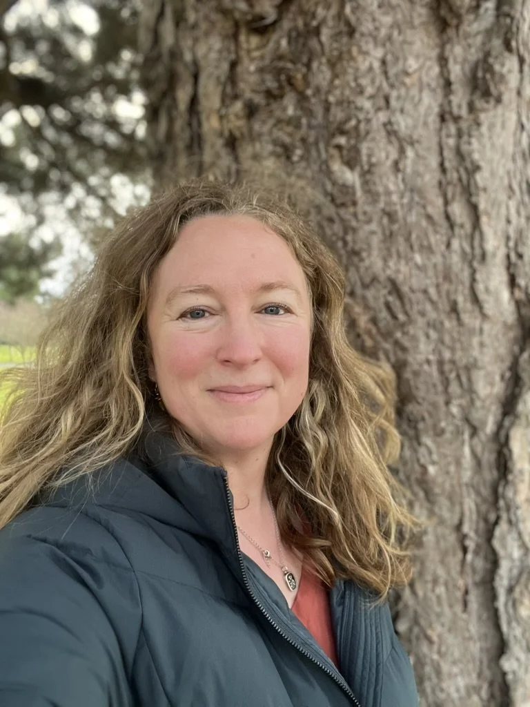 A woman with wavy, blonde hair taking a selfie outdoors next to a large tree trunk, wearing a dark puffer jacket and a maroon top.