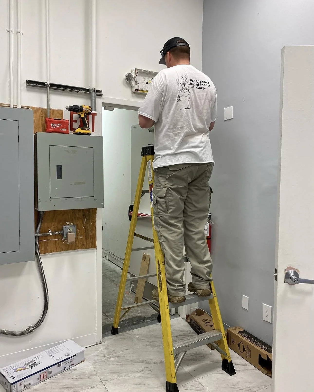 A worker standing on a yellow ladder, installing or repairing an emergency exit light fixture in a room. The worker is wearing a white T-shirt with black text on the back and beige work pants.