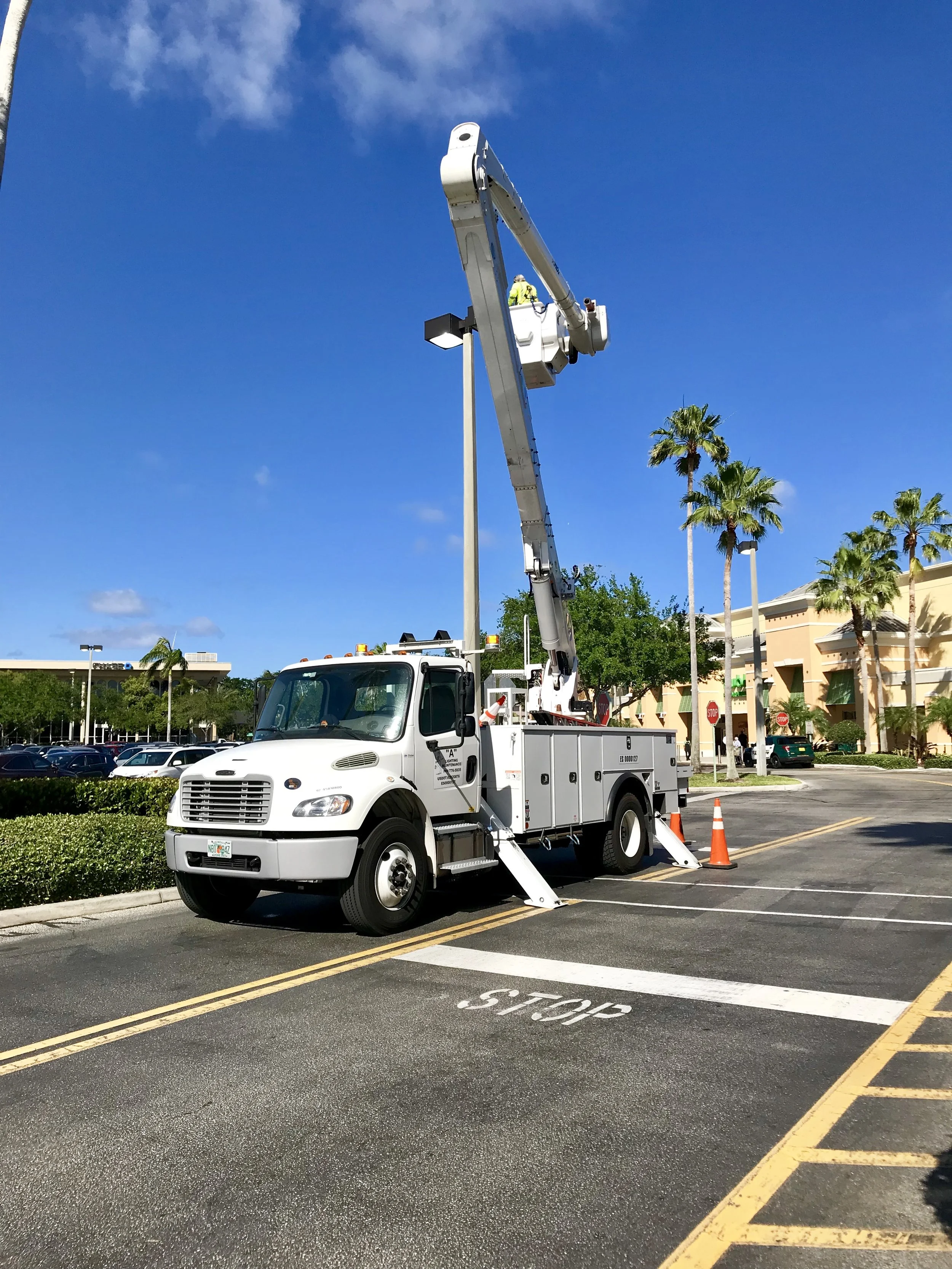 A utility truck with an extended boom lift working on a street in a shopping center parking lot. The truck is white with orange safety cones placed around it. In the background, there are palm trees, shopping centers, and a bright blue sky with some clouds.