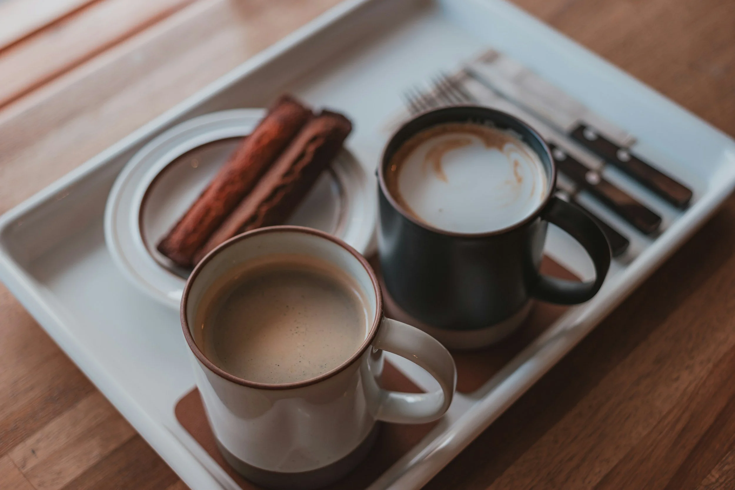 Two white mugs with coffee, a small cup with foam art, and two chocolate croissants on a white tray on a wooden table.