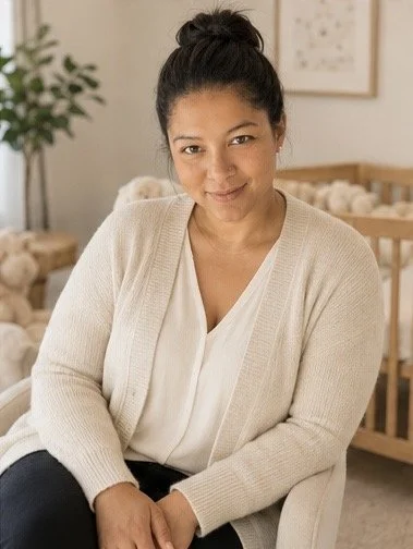 Portrait of a smiling woman with dark hair tied up in a bun, wearing a beige cardigan and white blouse, sitting in a cozy room with a crib and plant in the background.