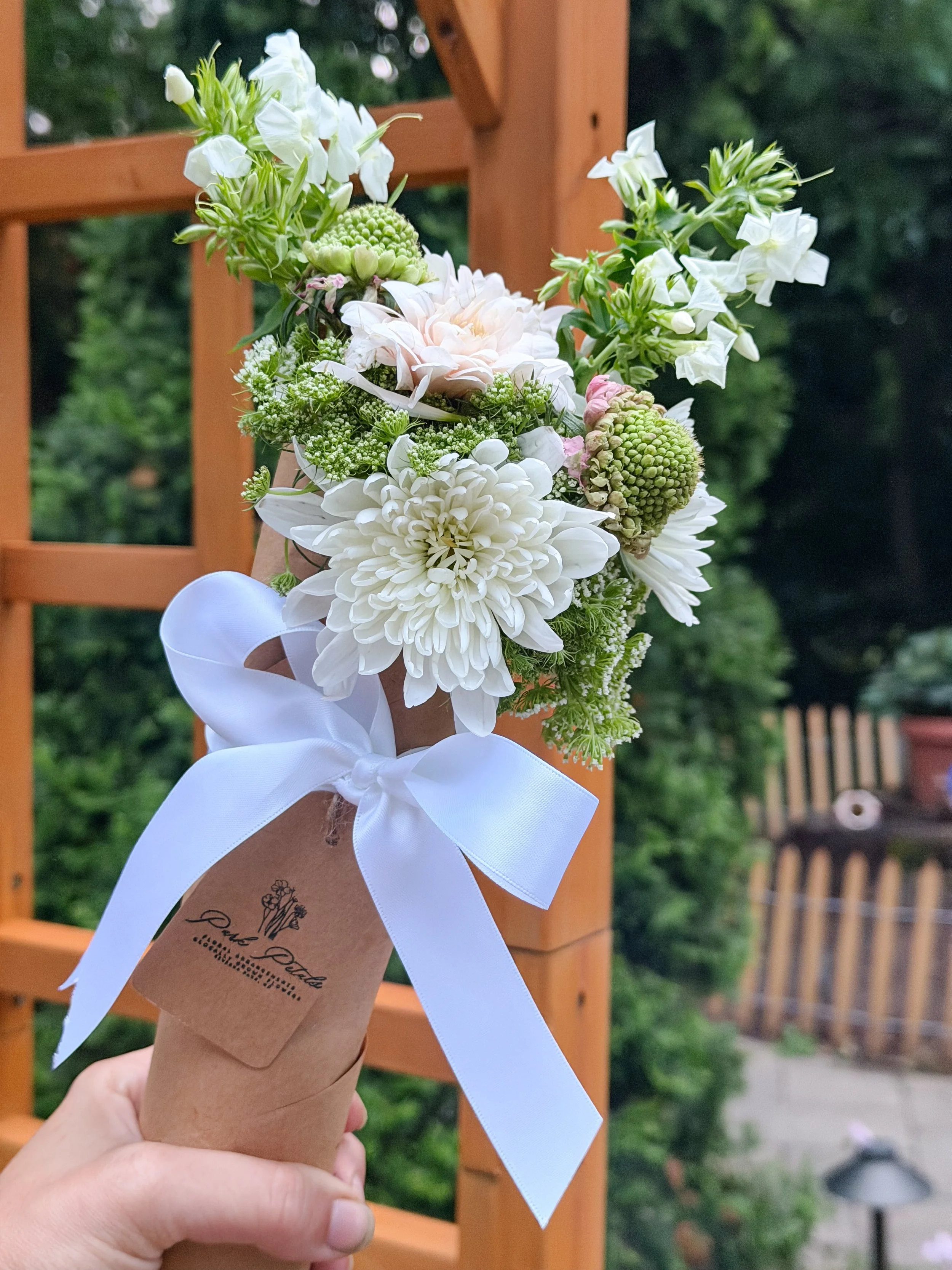 A hand holding a flower bouquet wrapped in brown paper with a white ribbon bow, set against a wooden trellis in an outdoor garden.