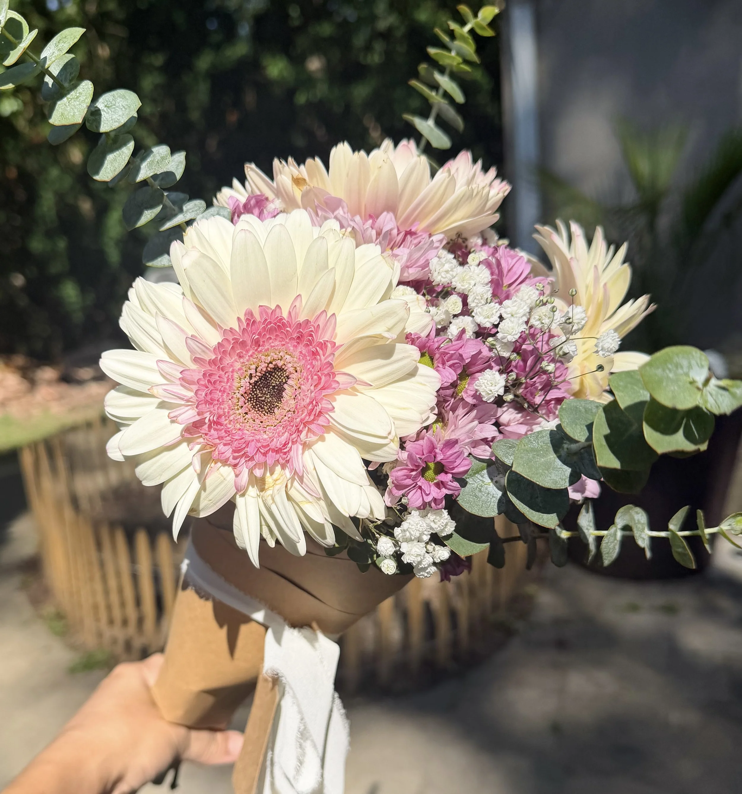 A person holds a bouquet of flowers with white petals and pink centers, surrounded by small white filler flowers and green leaves, against a blurred outdoor background.