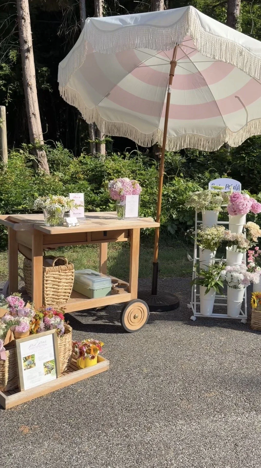 Outdoor flower stand under a large white parasol with floral arrangements in white vases and baskets, displayed on a wooden cart, chair, and shelves.