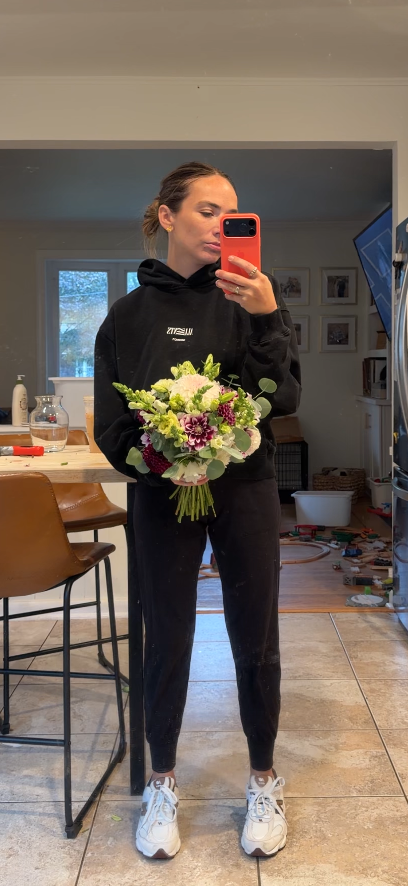 A woman in a black hoodie and leggings holds a bouquet of flowers and takes a mirror selfie in a kitchen with children’s toys on the floor.