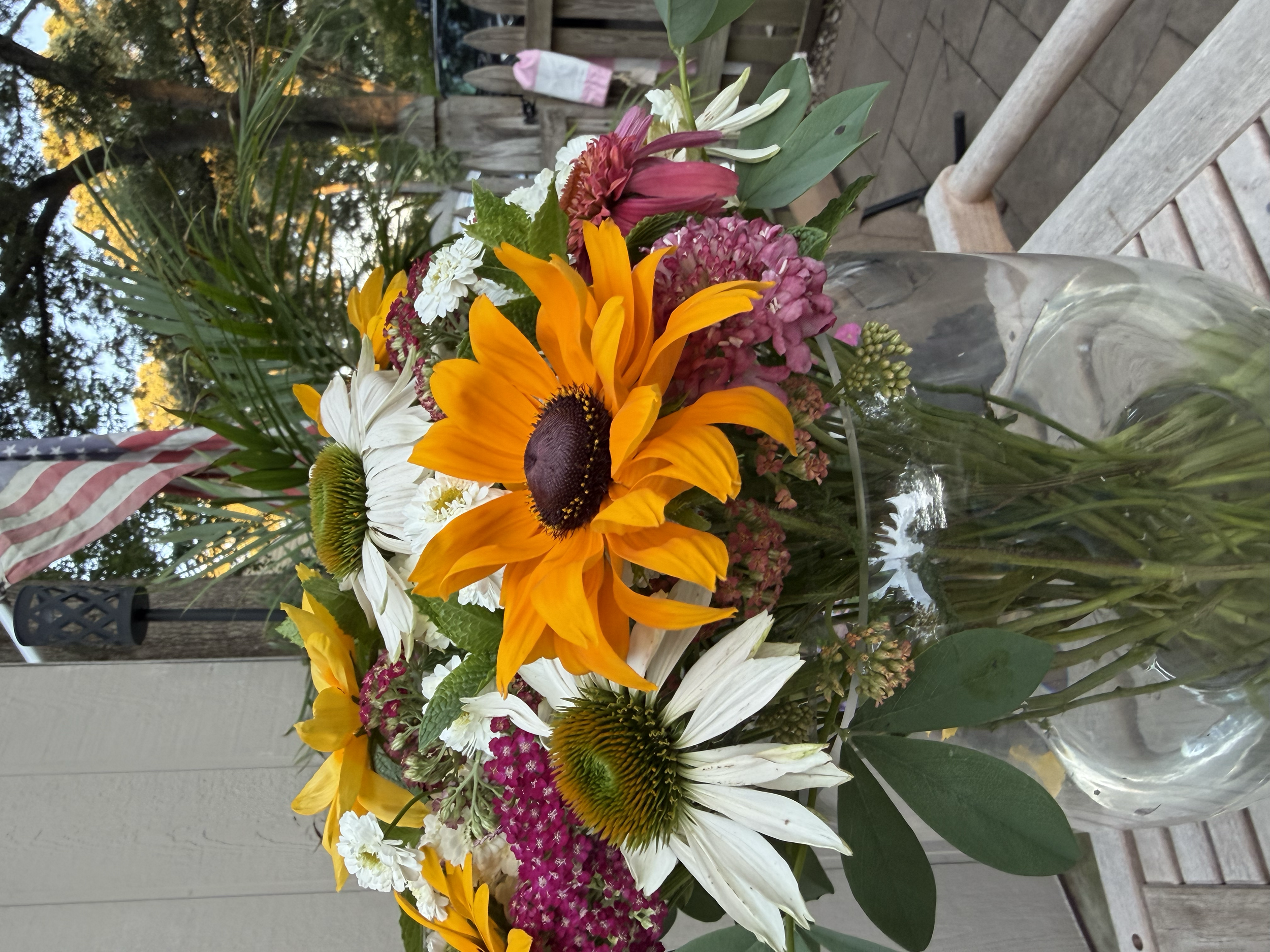 A colorful bouquet of flowers in a glass vase, including sunflowers, white daisies, pink and white carnations, and other mixed flowers, placed on a wooden surface outdoors with trees and a fence in the background.