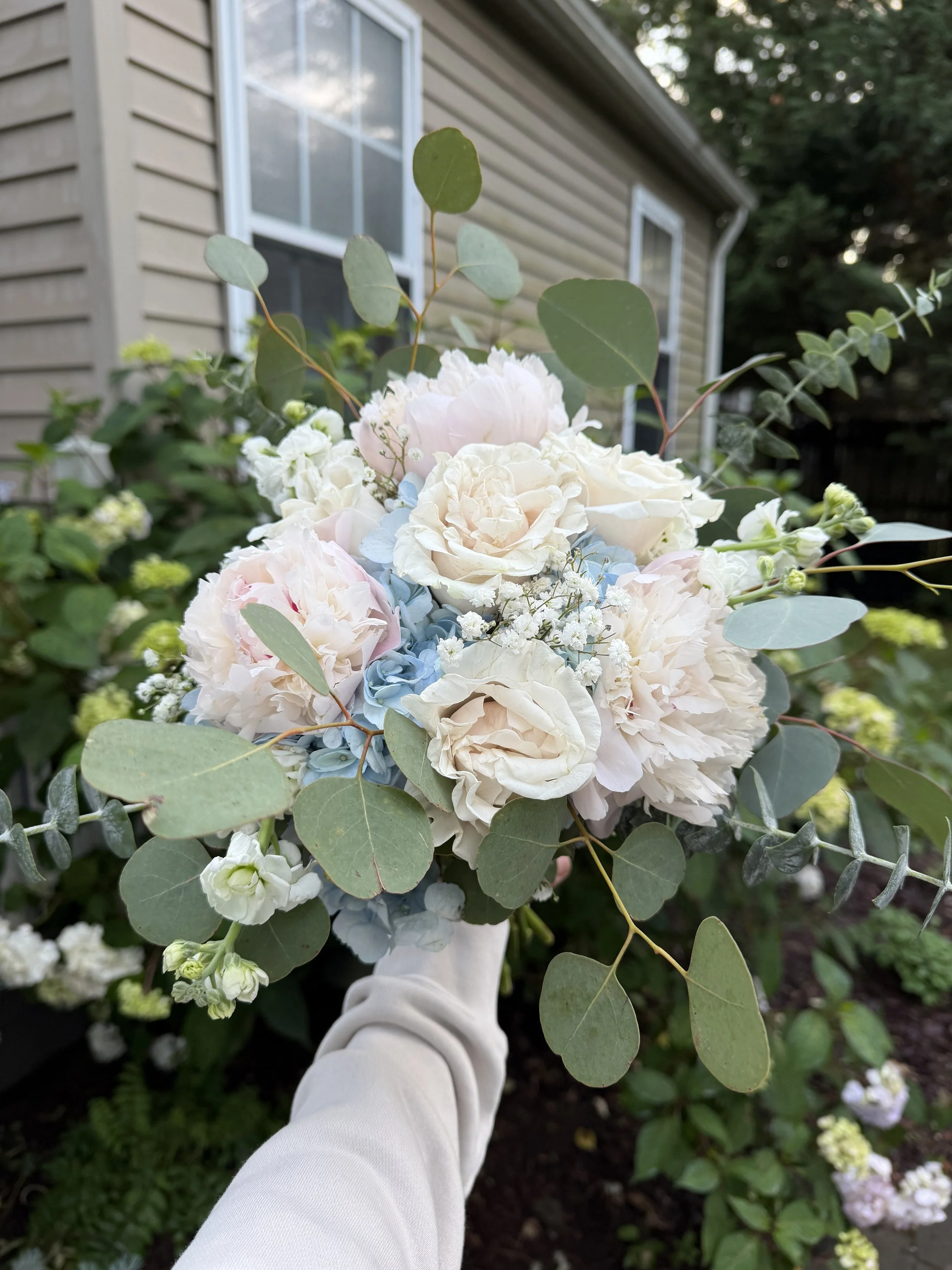 A person wearing a white long sleeve shirt holding a bouquet of white and pastel-colored hydrangeas and roses with green leaves against a house with siding and windows.