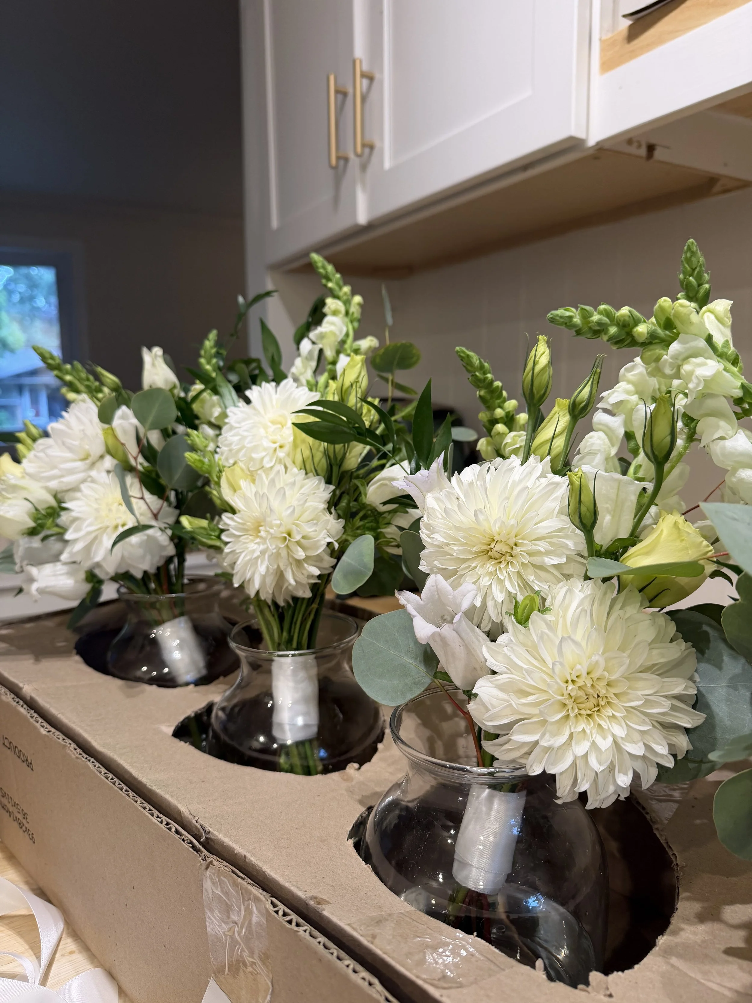 White and cream-colored floral bouquets in glass vases, placed on a cardboard box in a kitchen setting.