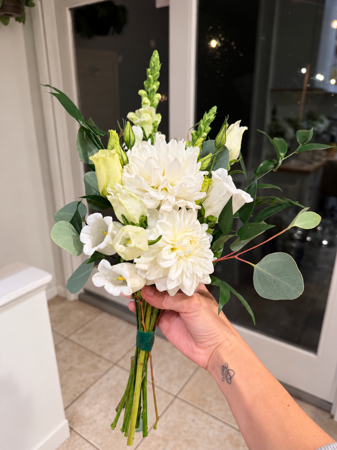 A hand holding a bouquet of white flowers and green leaves indoors near a glass door at night.