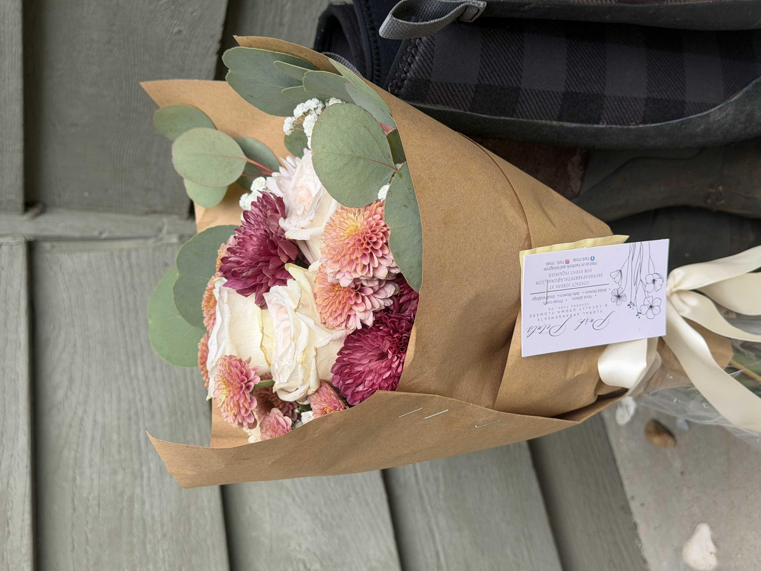 A bouquet of flowers wrapped in brown paper with a white ribbon, placed next to a black bag.