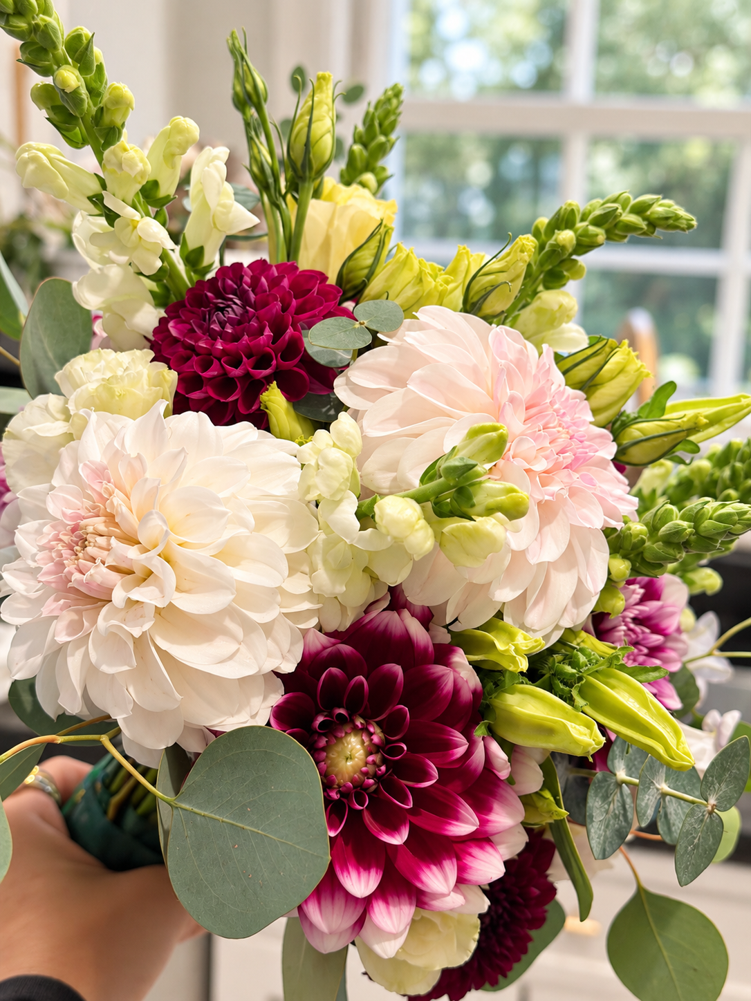 A bouquet of various flowers including dahlias, roses, and greenery in front of a window with sunlight.