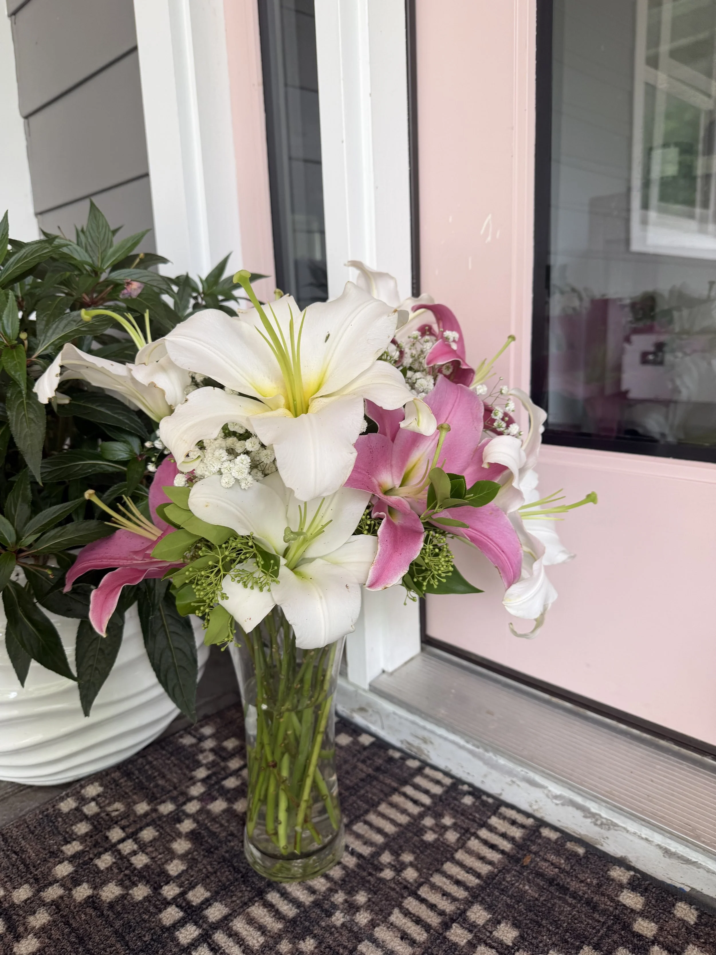 A glass vase filled with white, pink, and yellow lilies, baby's breath, and green leaves placed on a patterned carpet in front of a pink and gray house exterior window.