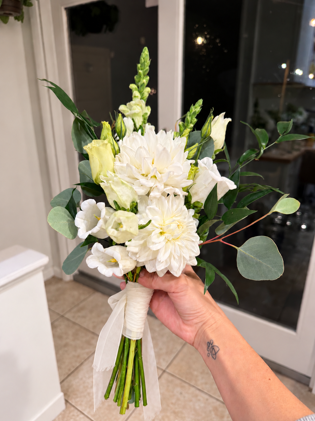 A hand holding a bouquet of white flowers with greenery, wrapped with white ribbon, indoors near a glass door.