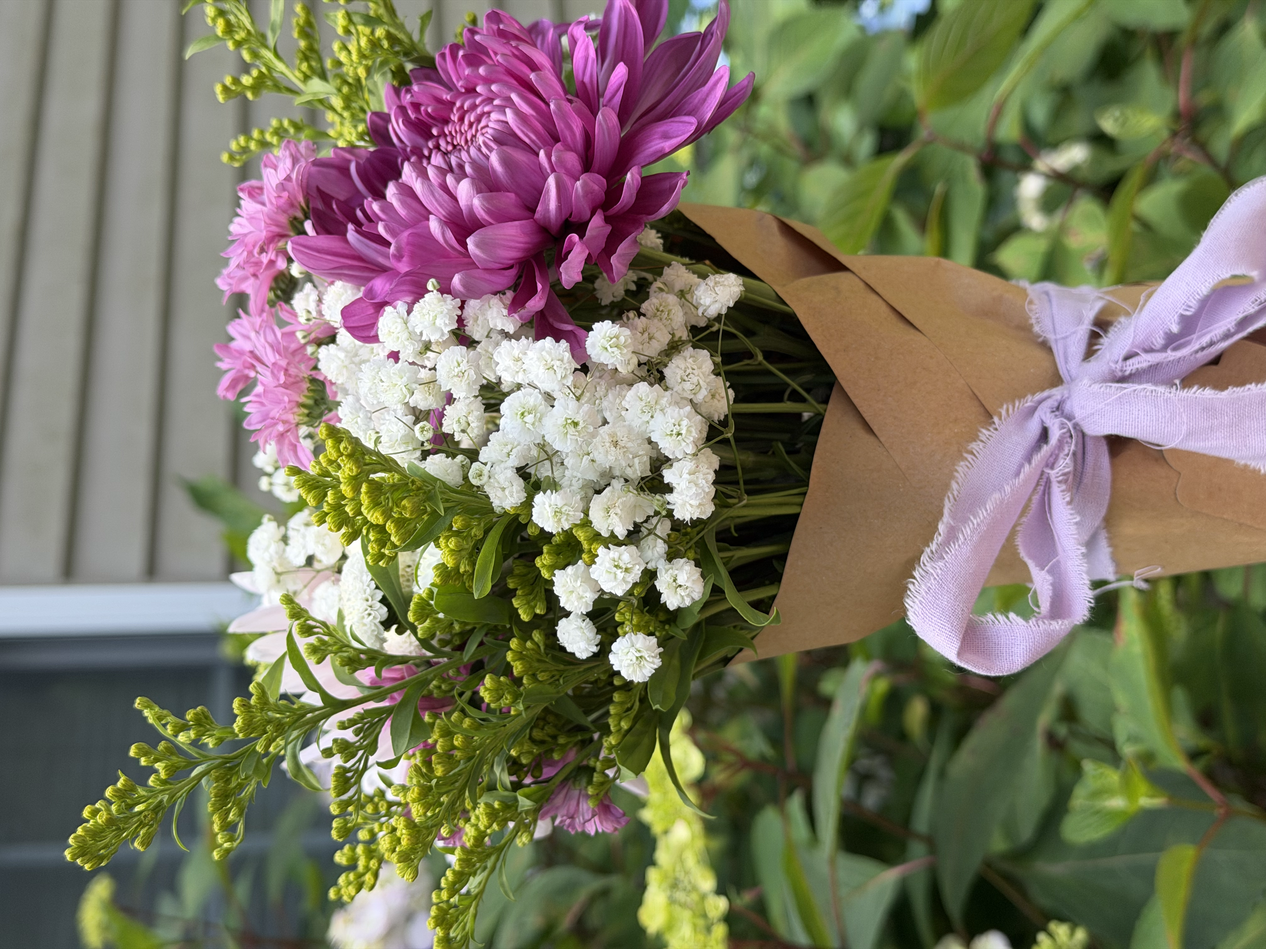 A bouquet of pink and white flowers wrapped in brown paper and tied with a lavender ribbon. The bouquet includes large pink chrysanthemums, small white blooms, and green foliage.