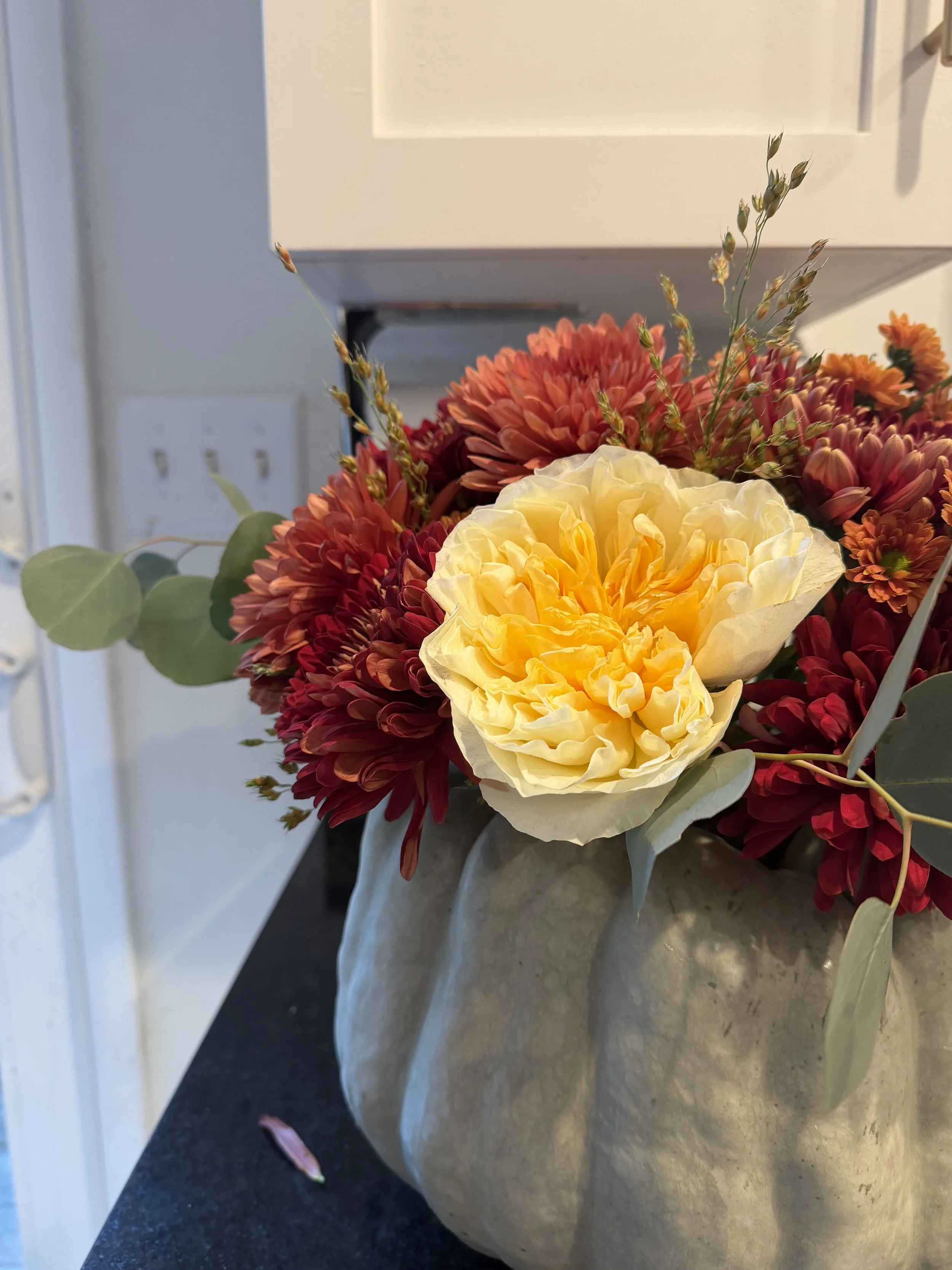 A bouquet of orange, red, and yellow flowers in a beige ceramic vase on a gray countertop, with a white wall and electrical outlet in the background.