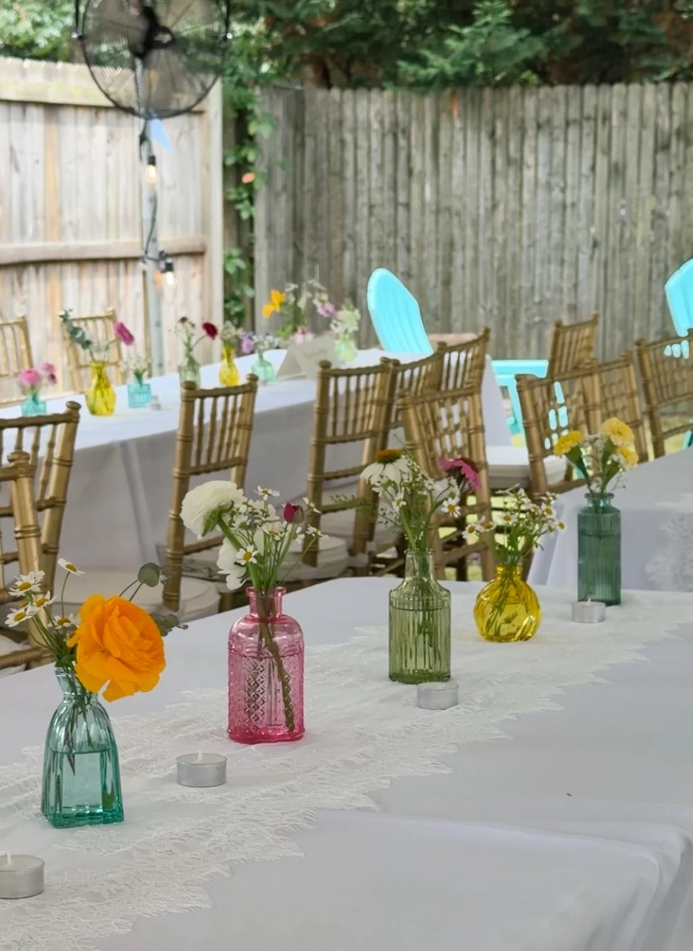 Outdoor celebration with decorated tables, floral centerpieces in colorful glass vases, tealight candles, wooden chairs, and string lights in a backyard with a wooden fence and trees.