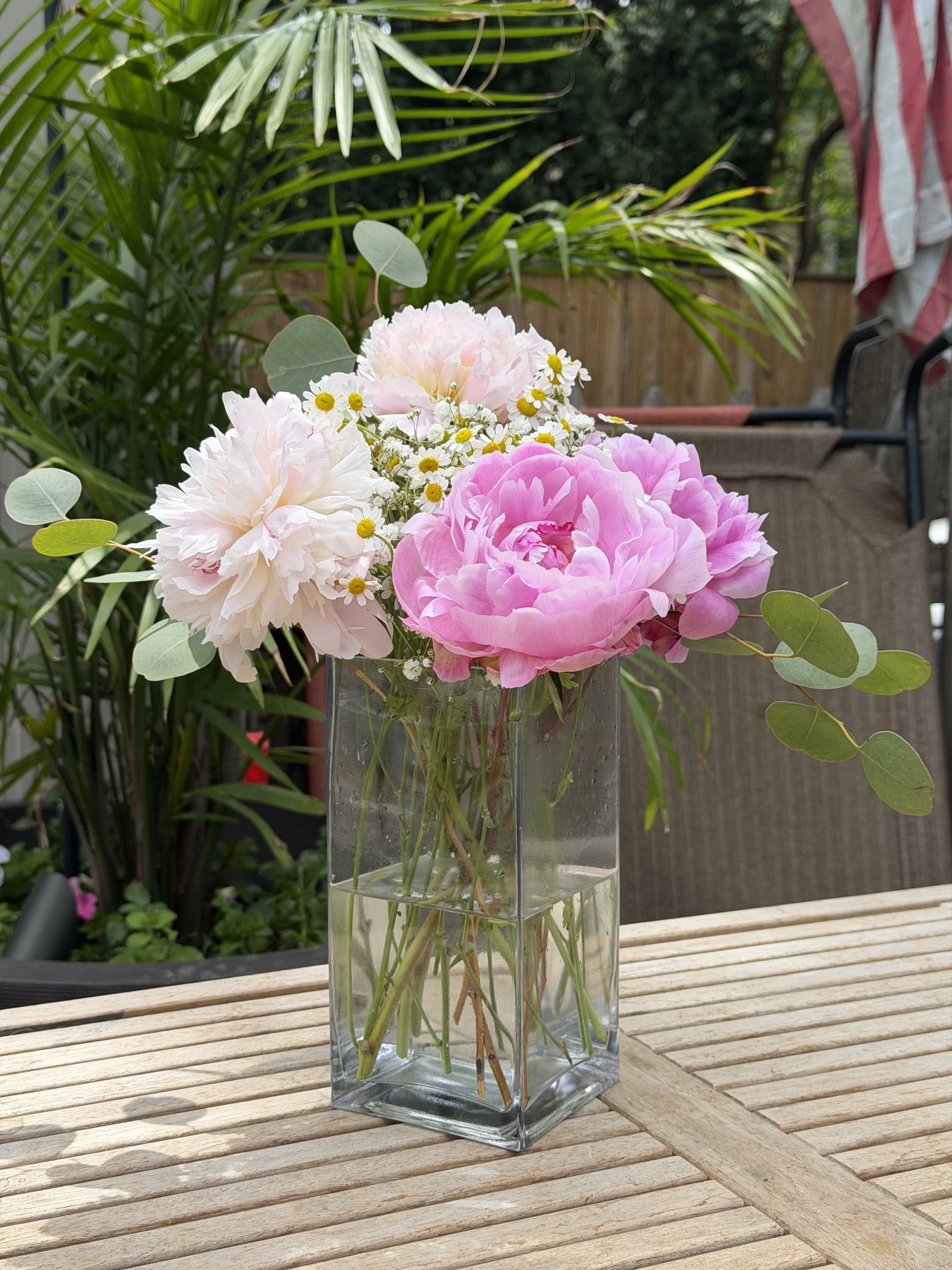 A glass vase with pink and white peonies and small white daisies on a wooden table in a garden setting.