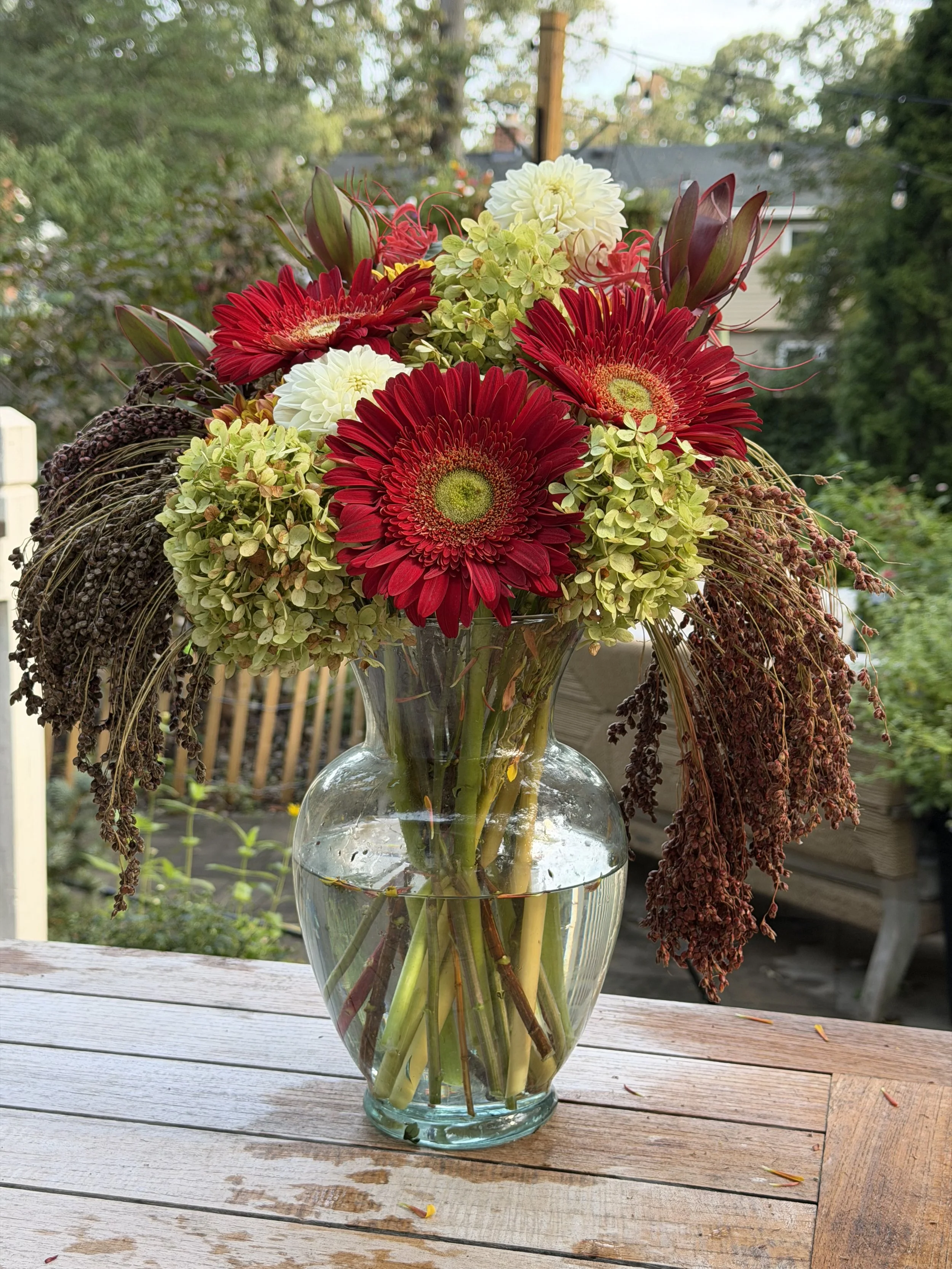 A glass vase filled with water and flowers on a wooden table outdoors. The bouquet includes red gerbera daisies, white dahlias, green hydrangeas, and brownish dried foliage. The background shows trees and a house with string lights.
