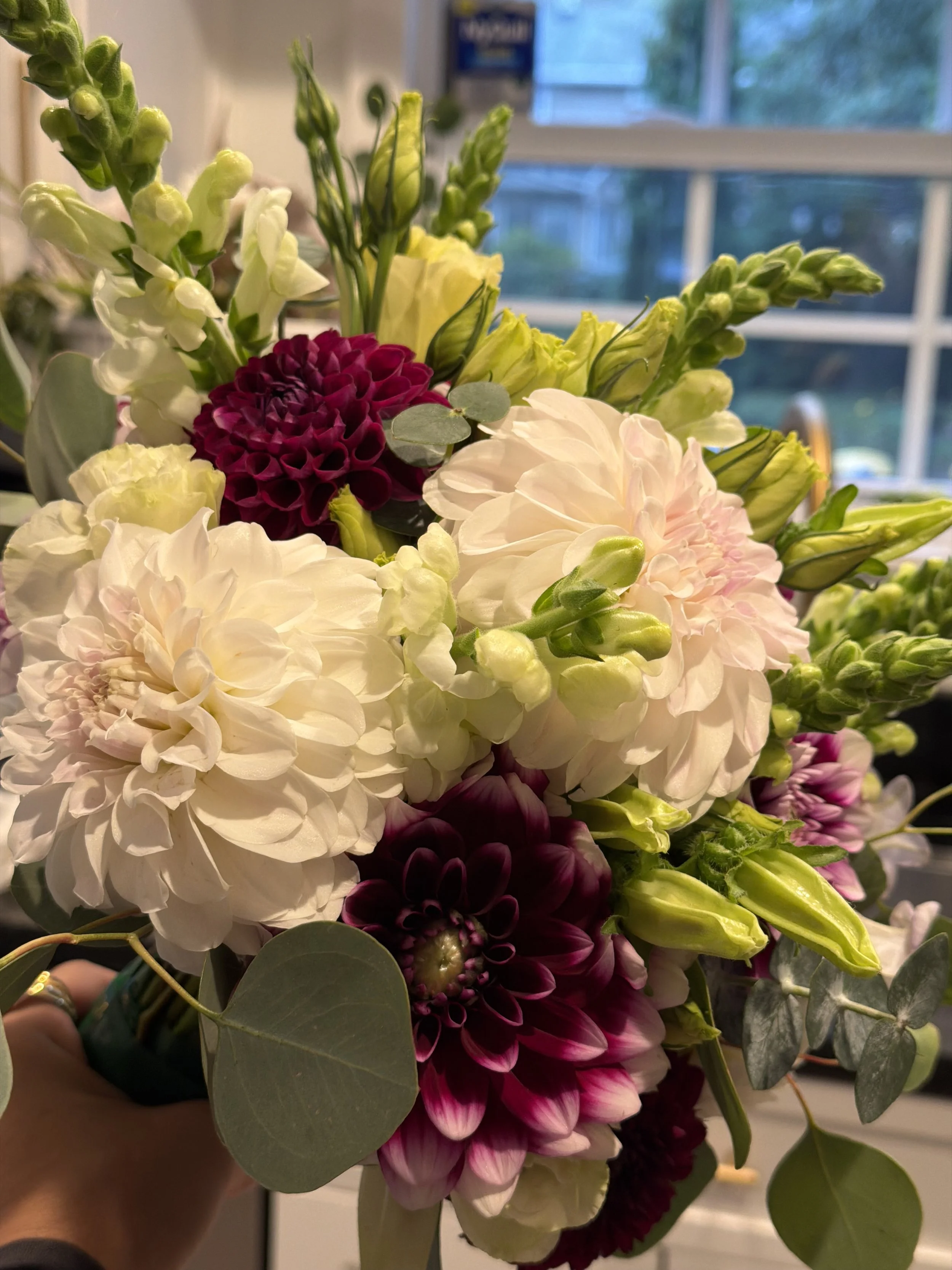 A bouquet of mixed flowers including white, deep burgundy, and pink flowers, with green leaves and buds, held by a person.