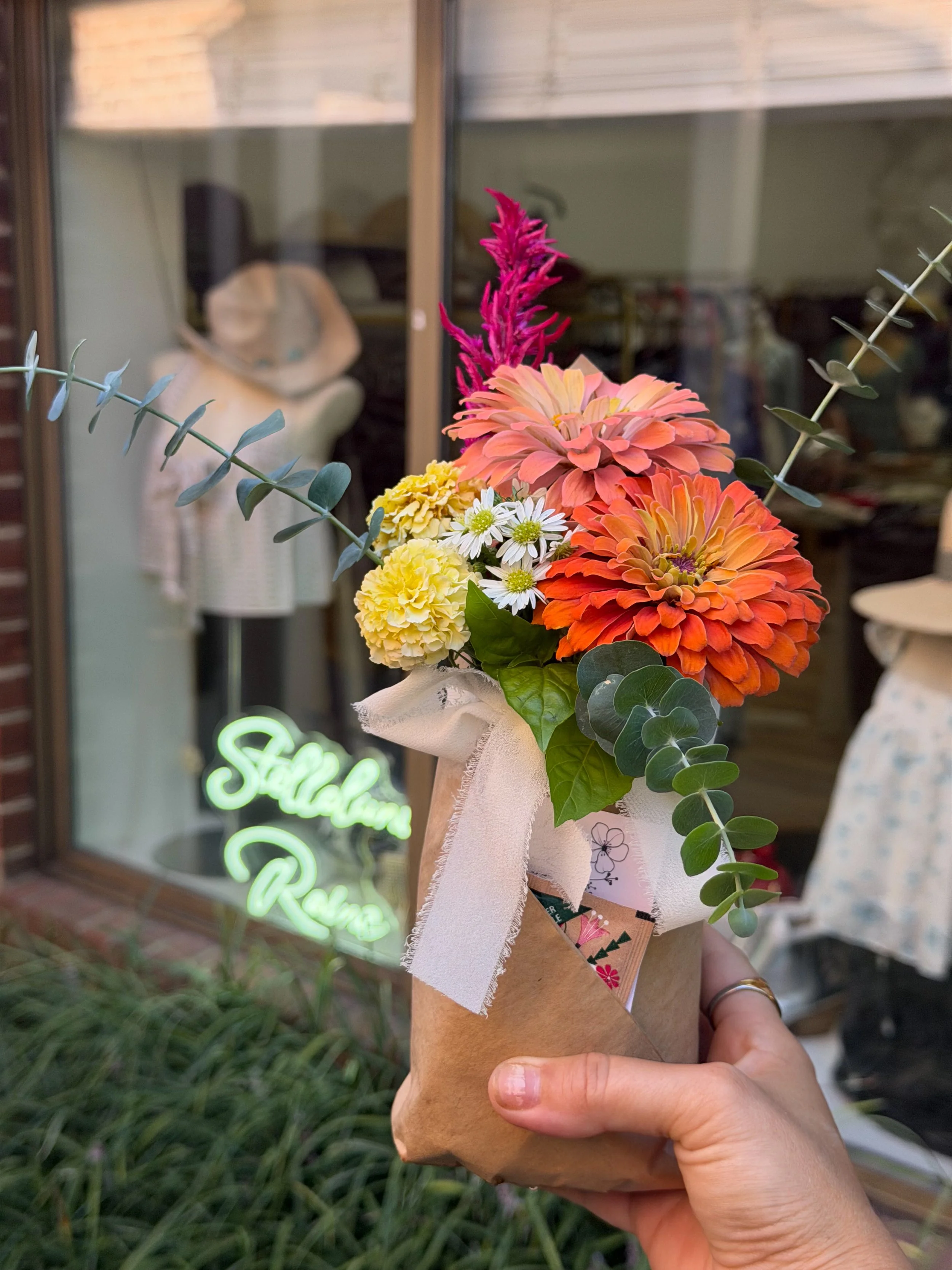 Hand holding a bouquet of colorful flowers wrapped in brown paper, with a blurred cafe window and greenery in the background.