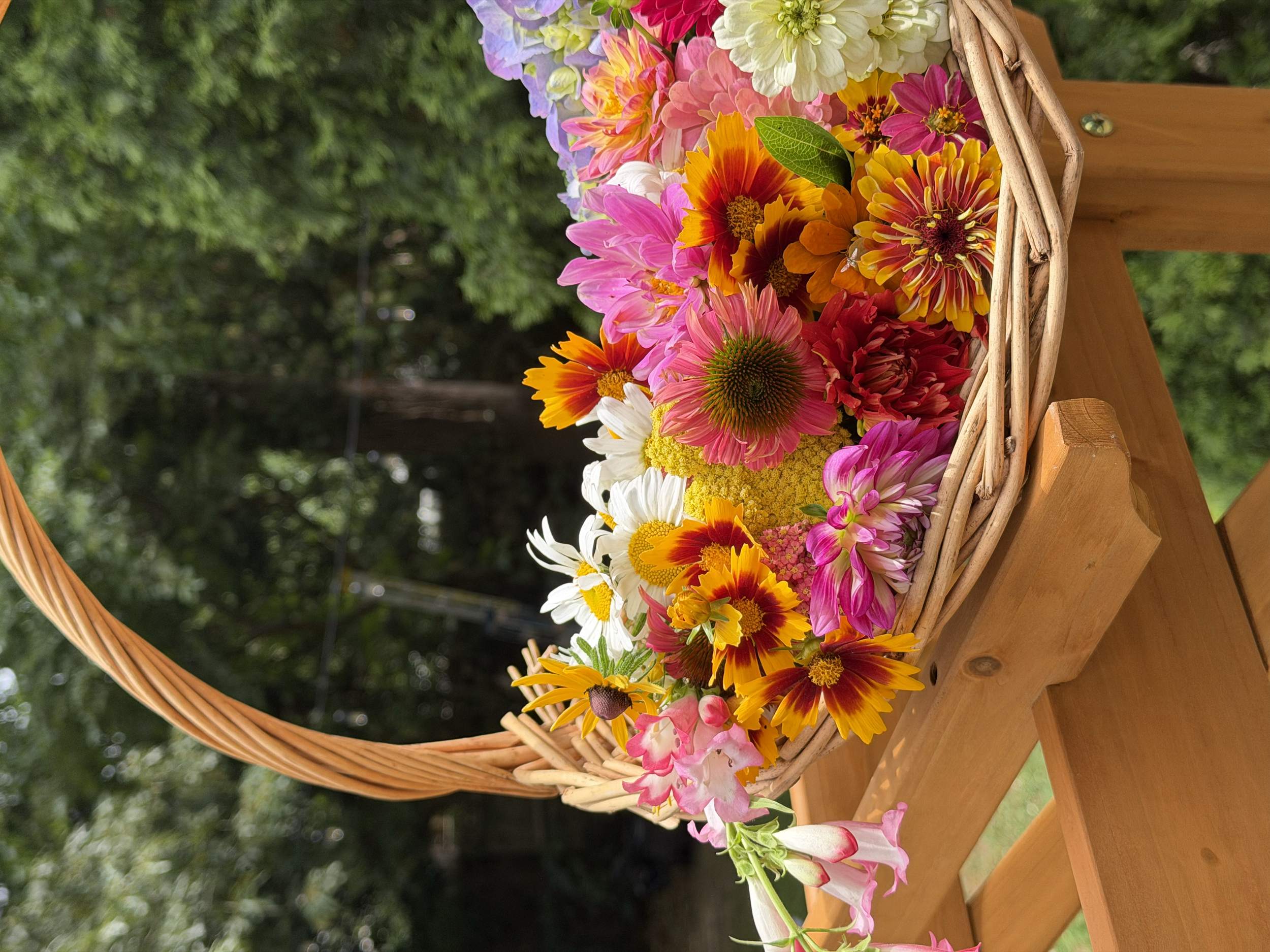 Colorful bouquet of various flowers in a wicker basket on a wooden table outdoors, with green foliage in the background.