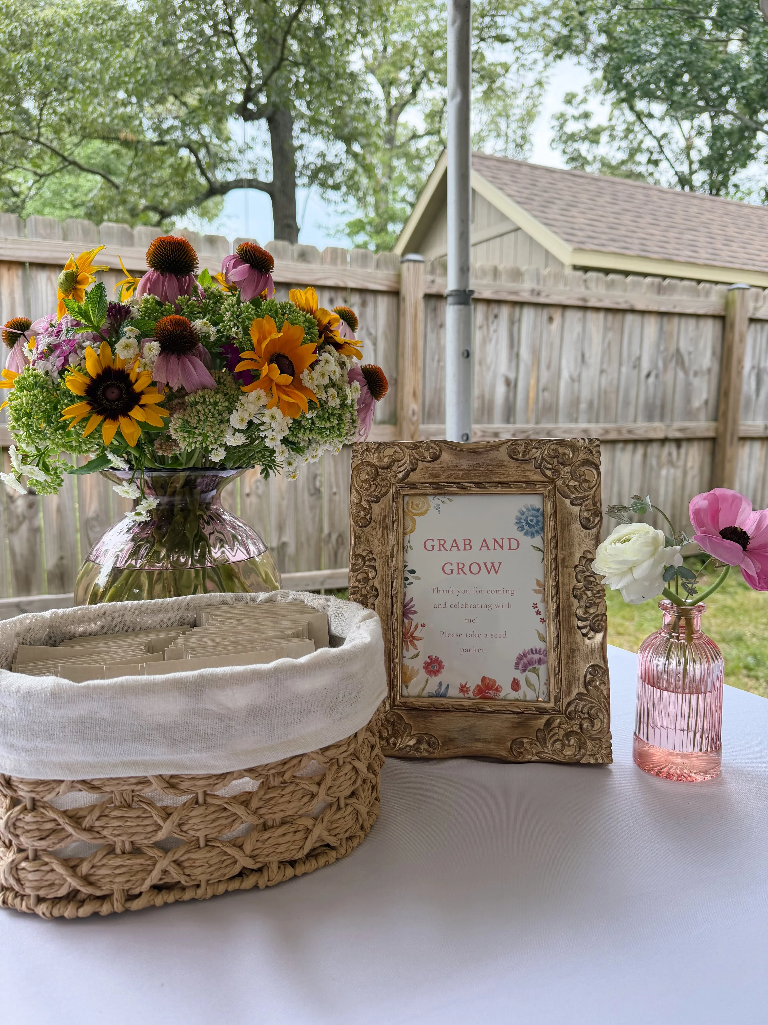 A large bouquet of mixed flowers in a glass vase on a table with a sign that reads 'Grab and Grow' and a wicker basket with seed packets inside, set outside with a wooden fence and trees in the background.