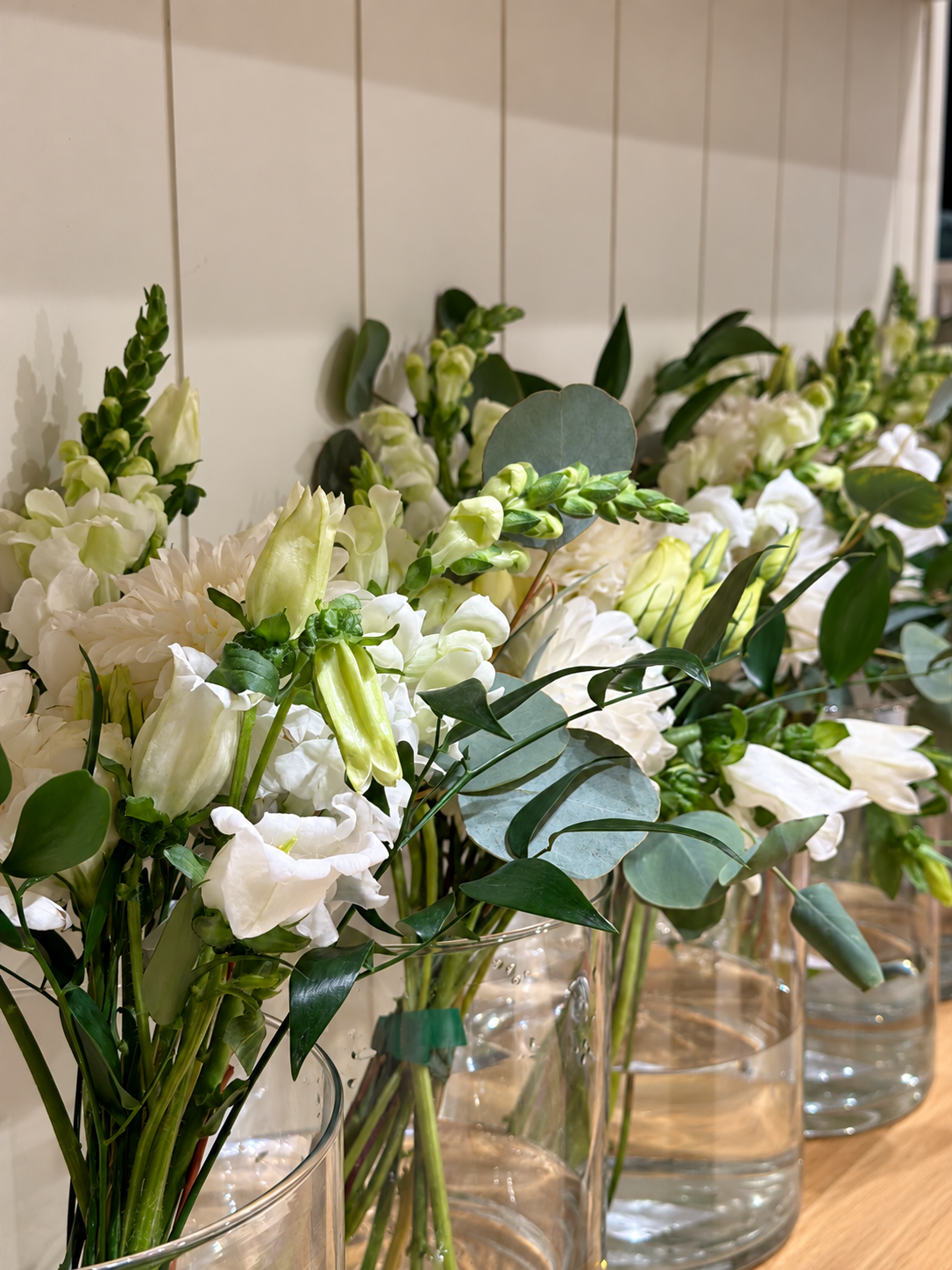 Several glass vases filled with white flowers and green foliage on a wooden surface, set against a white tiled wall.