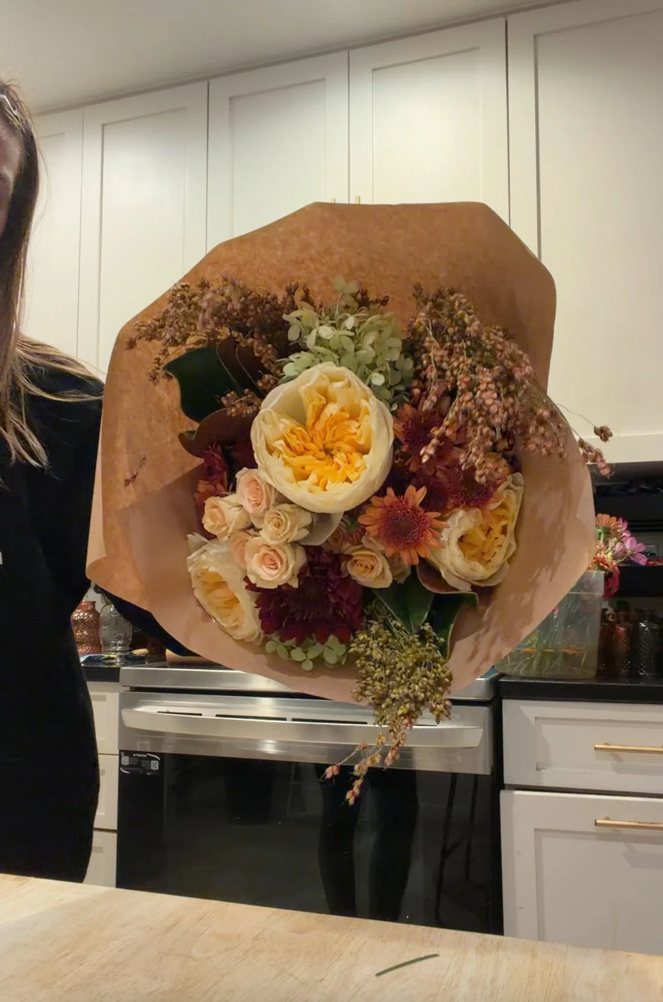 A large bouquet of flowers with cream, peach, and burgundy colors, wrapped in brown paper, being held by a person in a black top, in a kitchen with white cabinets and a black oven.