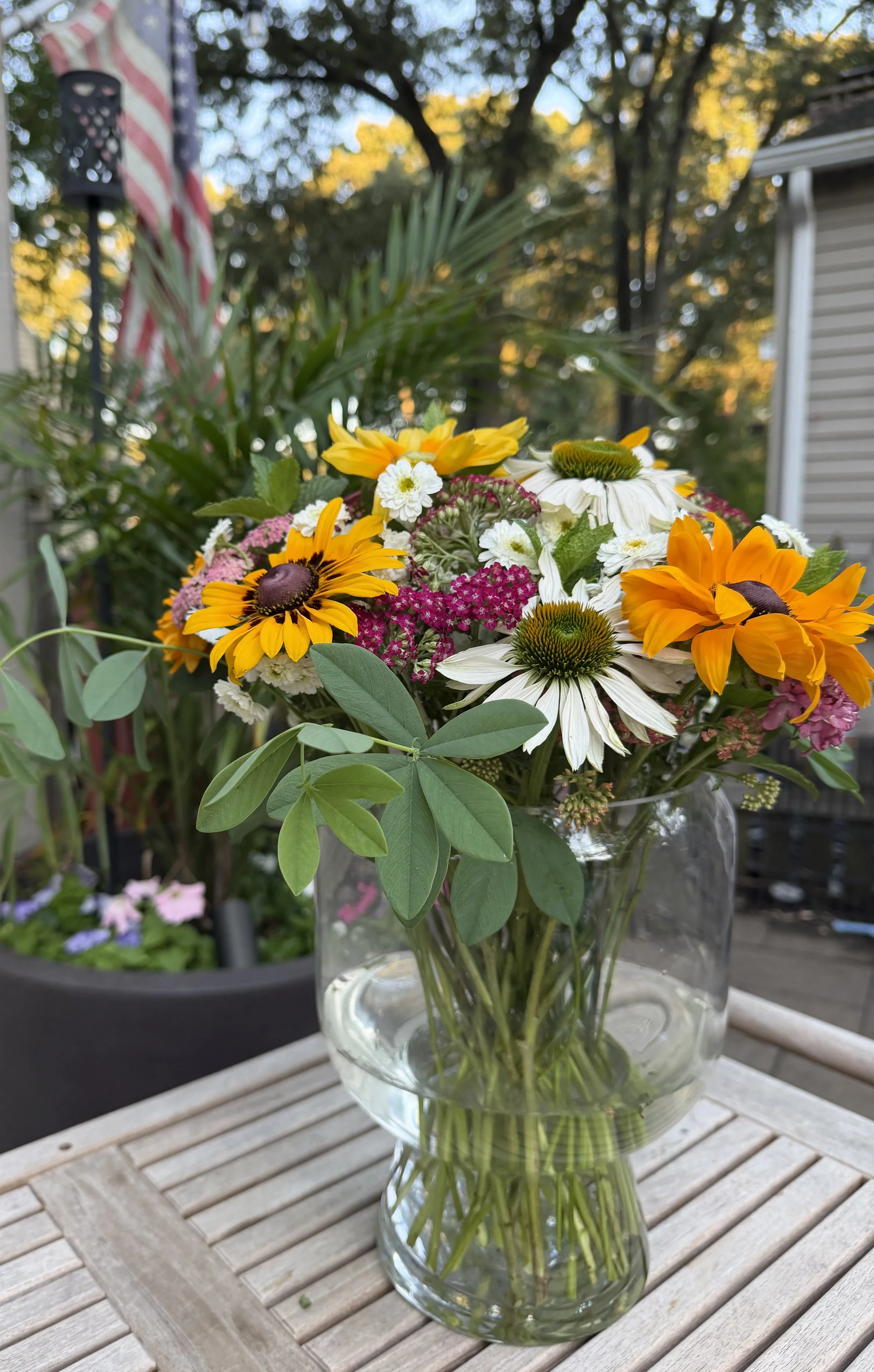 A glass vase filled with a colorful bouquet of flowers including white daisies, yellow black-eyed Susans, and pink and white flowers, placed on a wooden table outdoors during daytime.