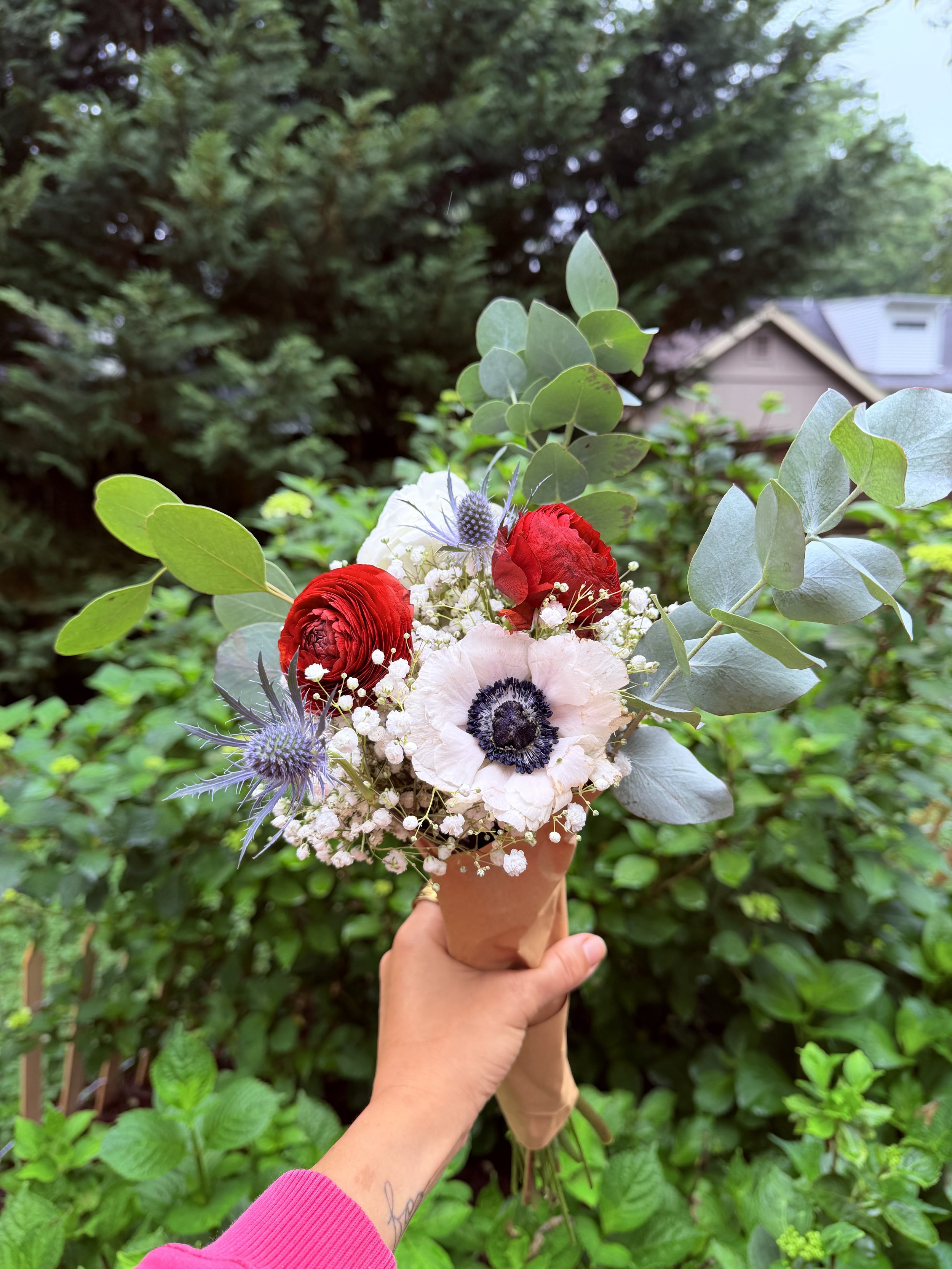 Person holding a bouquet of mixed flowers including red, white, and purple blooms against a background of green foliage and trees.