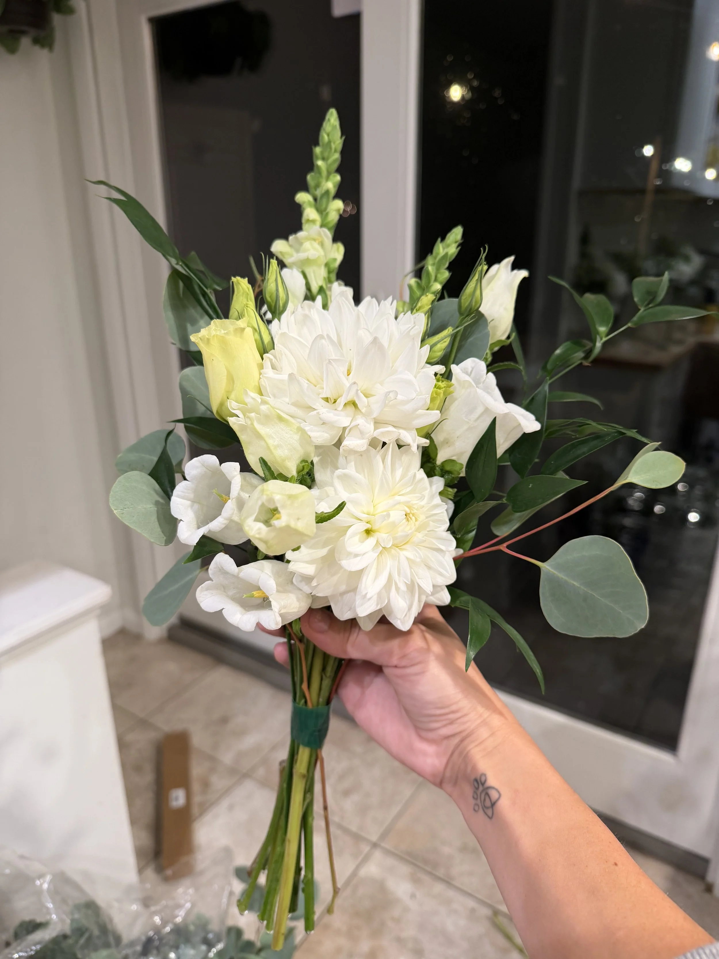 A person holding a bouquet of white and yellow flowers with green leaves inside a house near a window.