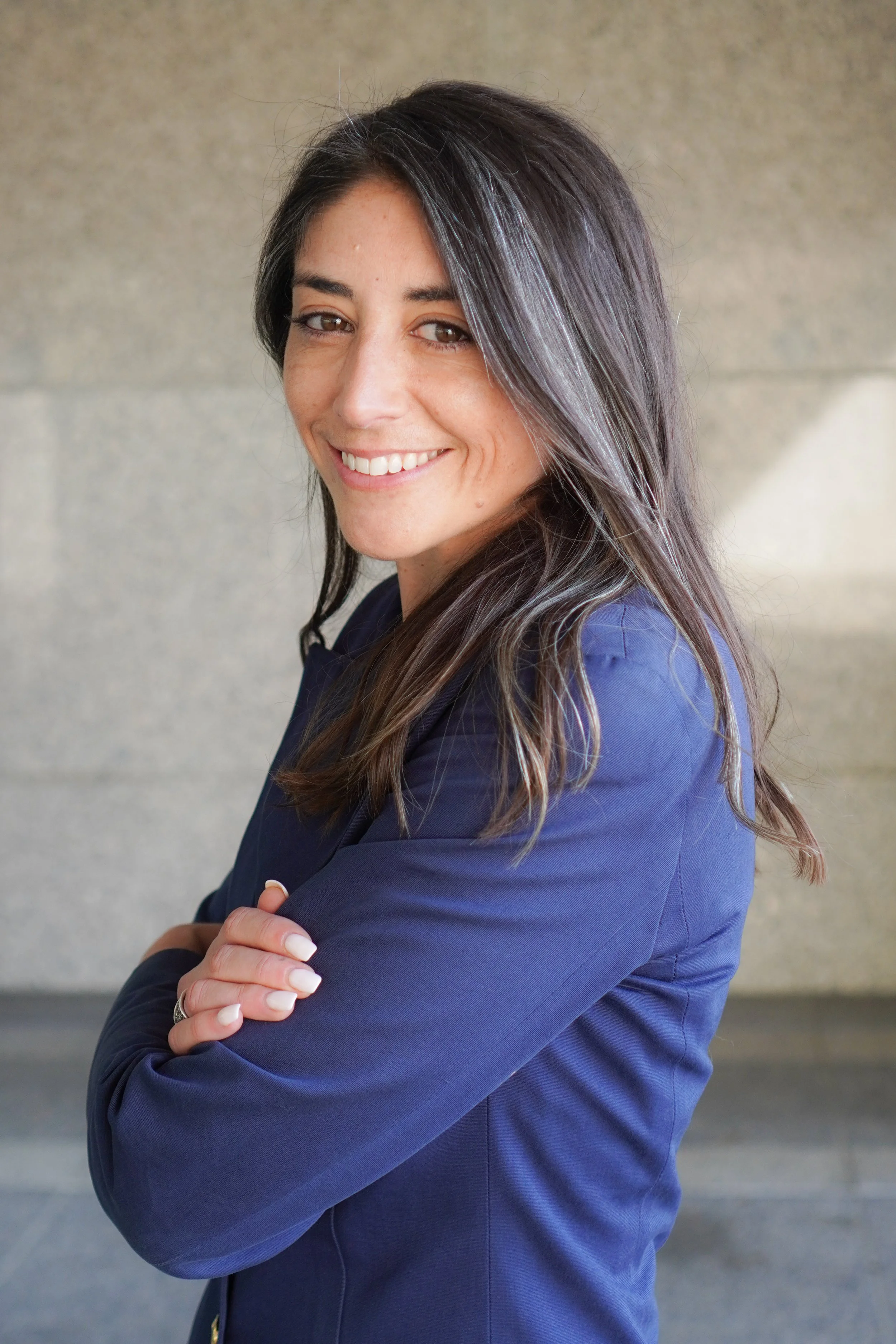 A woman with dark brown hair and brown eyes, smiling, wearing a navy blazer, standing with arms crossed against a neutral background.