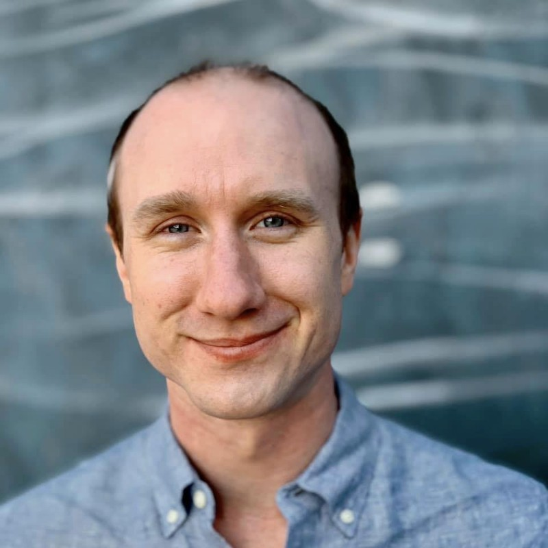 A man with a friendly smile, light skin, and short brown hair, wearing a light blue collared shirt, standing outdoors near a body of water.