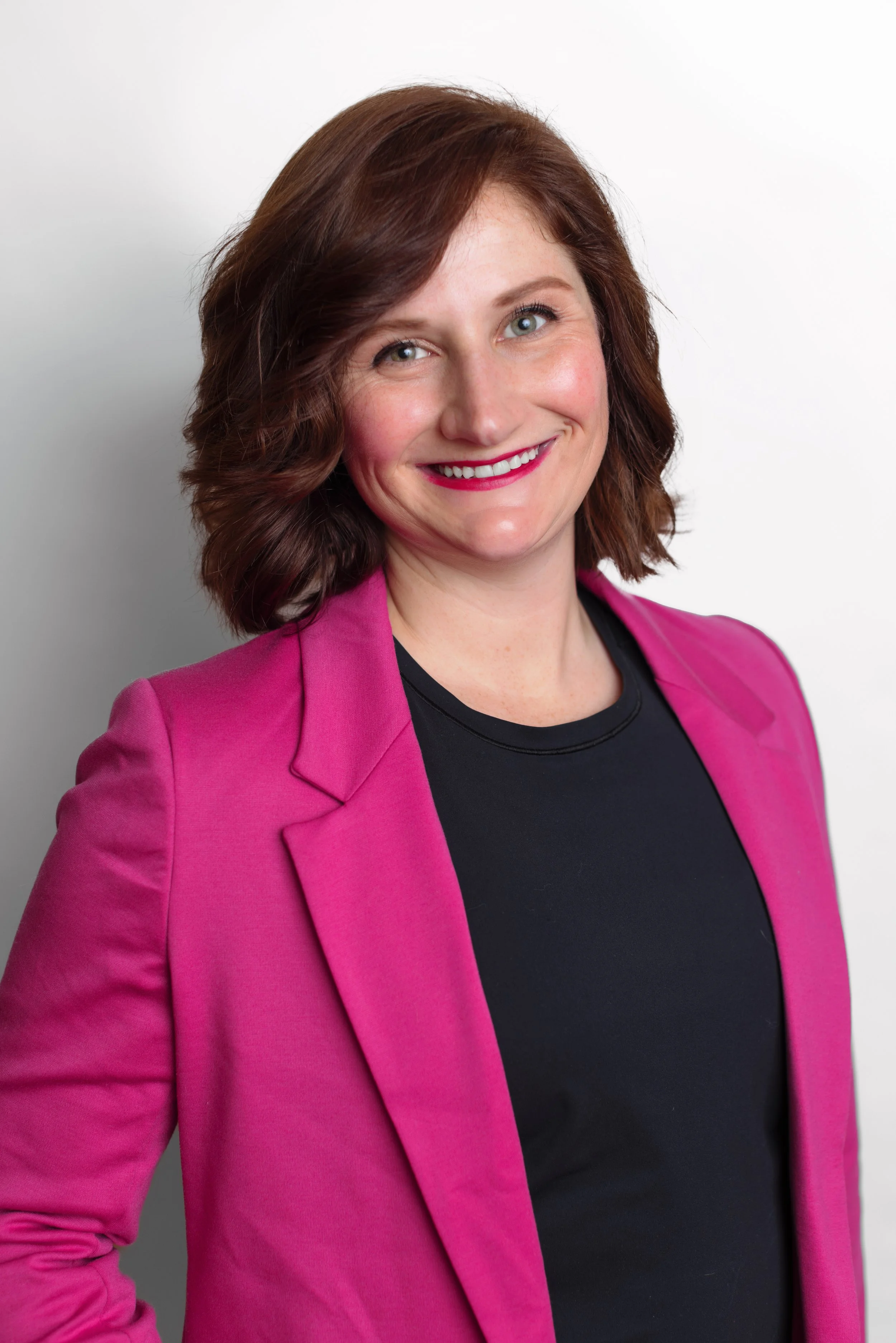 A woman with shoulder-length brown hair, smiling, wearing a bright pink blazer over a black top, against a plain white background.
